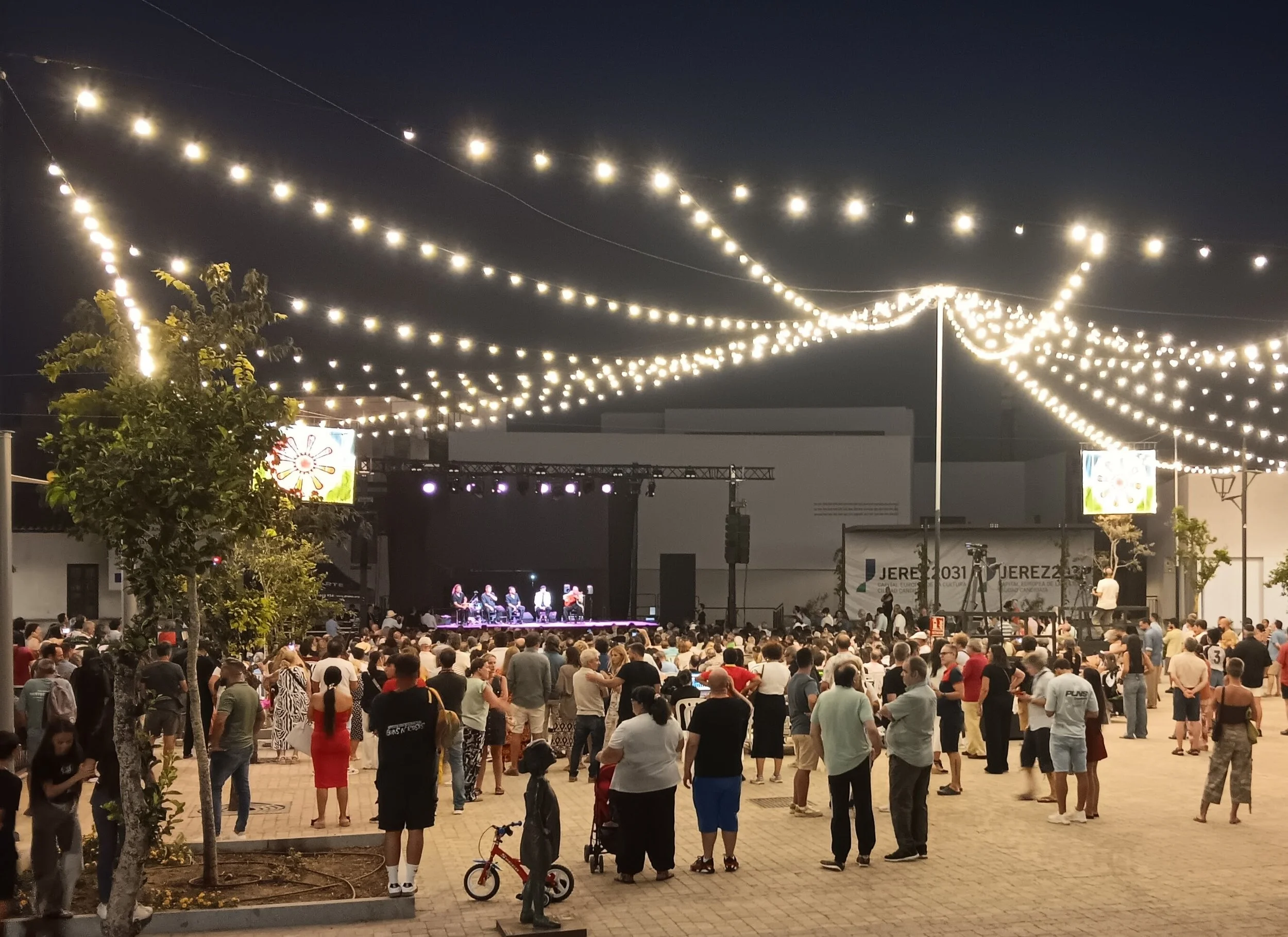 An outdoor concert with hundreds of people in a square watching performers on a stage. Above is an array of suspended lights.