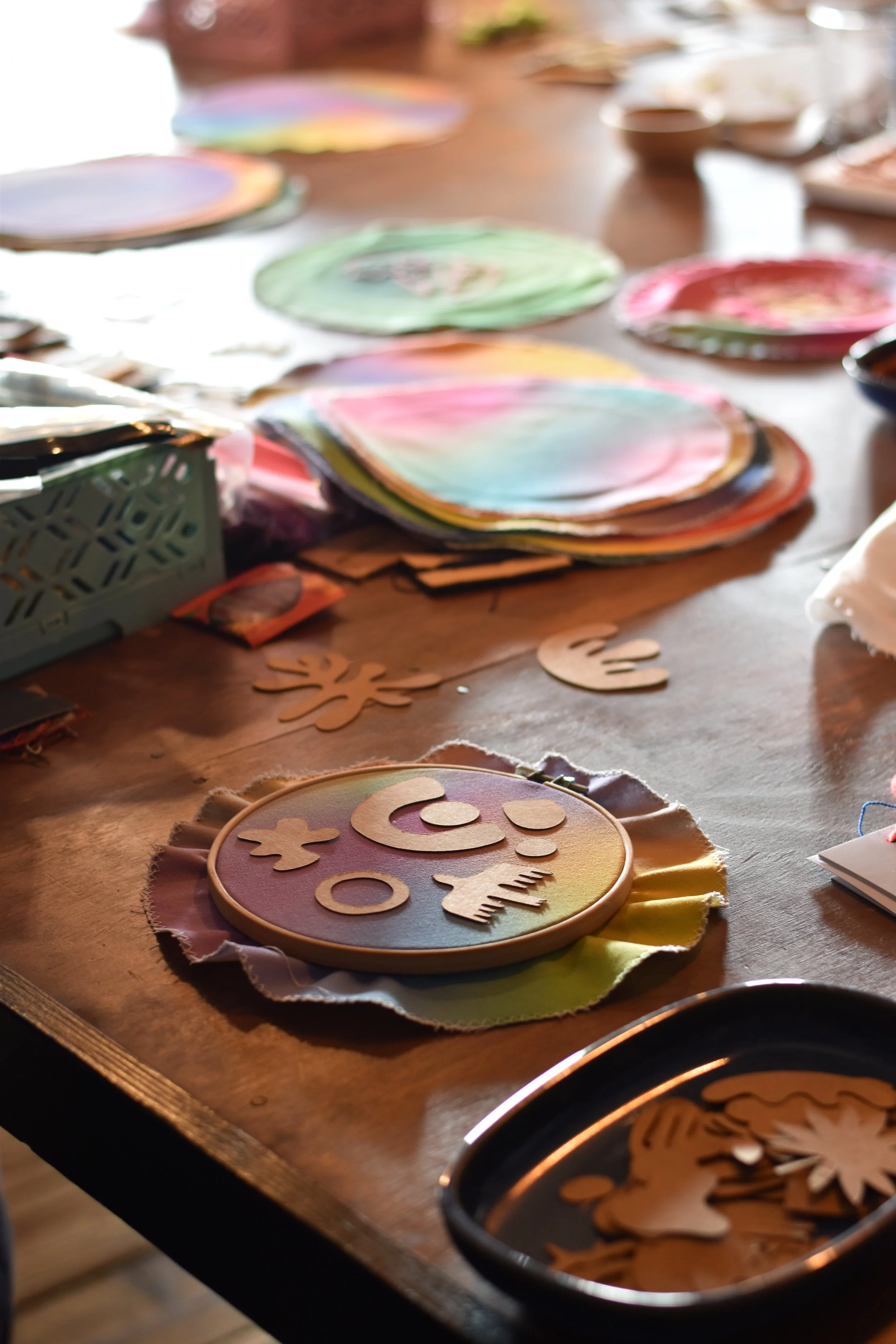Colorful paper plates and craft materials spread across a wooden table, with a prominently placed rainbow-colored embroidery hoop surrounded by wooden cutouts of various shapes.