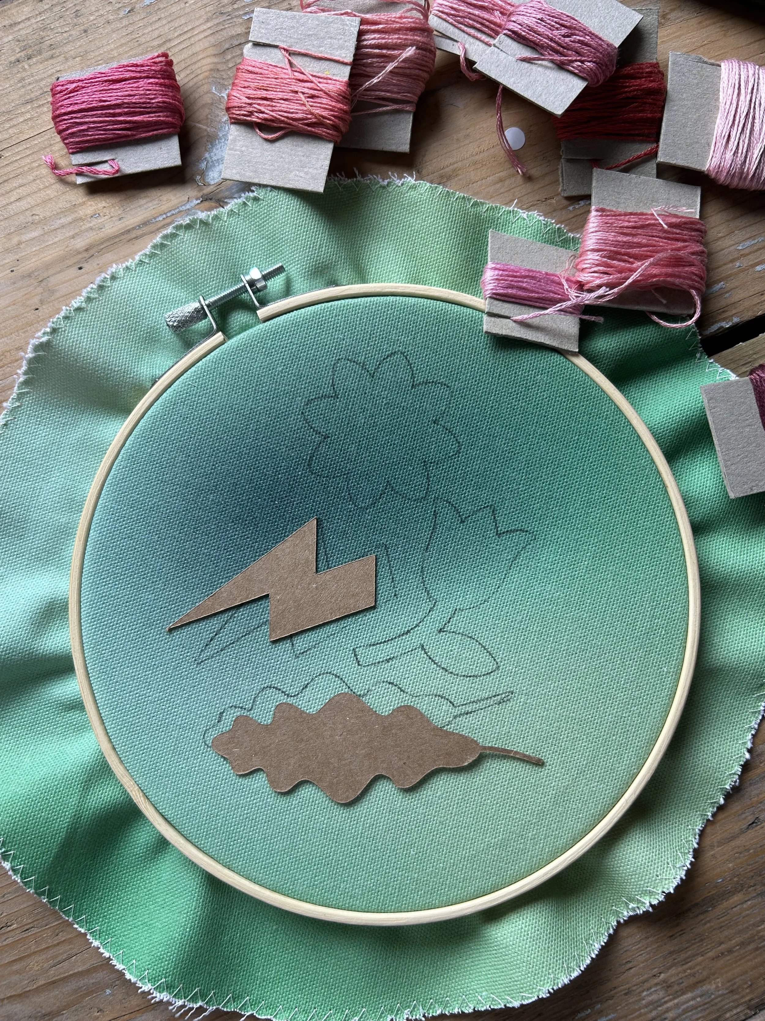 A green quilting hoop with some rough sketches of flowers, along with cutout cardboard shapes, a lightning bolt, and a cloud shape on a green fabric. Several spools of pink embroidery thread are arranged around the hoop on a wooden surface.