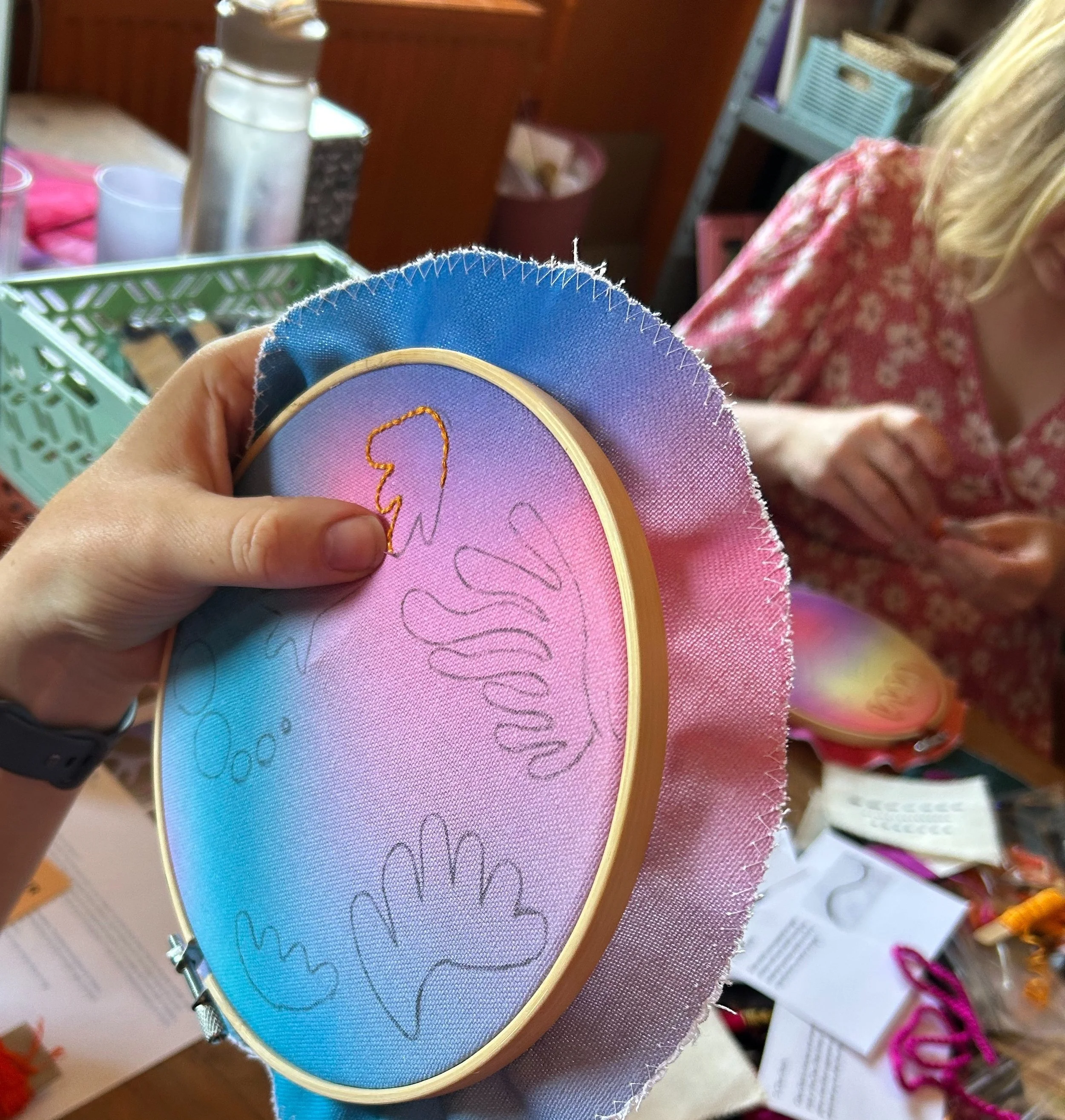 Close-up of an embroidery hoop with a pink and blue gradient fabric featuring an outline of a unicorn and clouds. A person is holding the hoop, and another person is working on a similar embroidery project in the background.