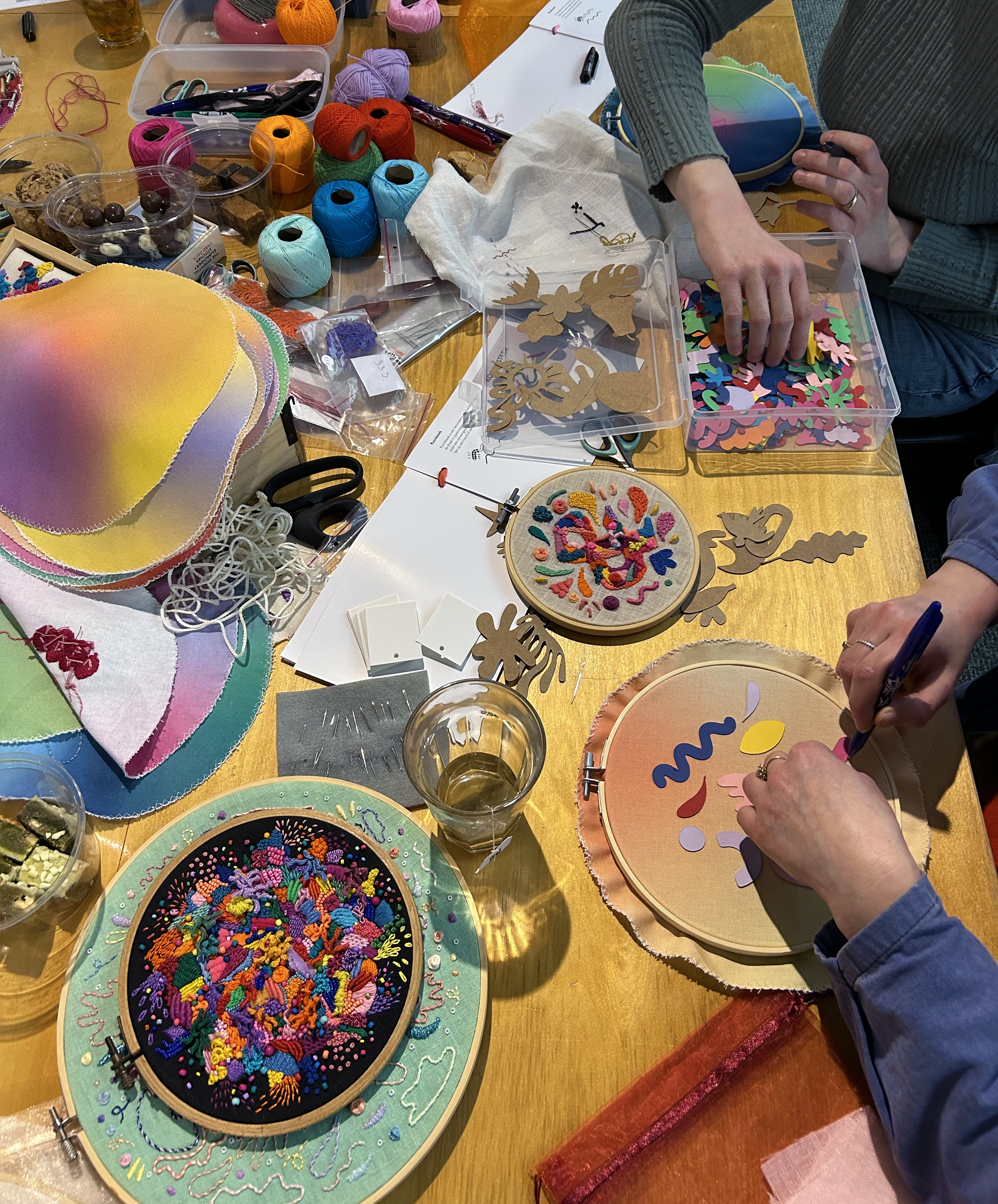 People making crafts with embroidery hoops, colorful fabric, cross-stitch, felt, and sewing supplies on a wooden table.