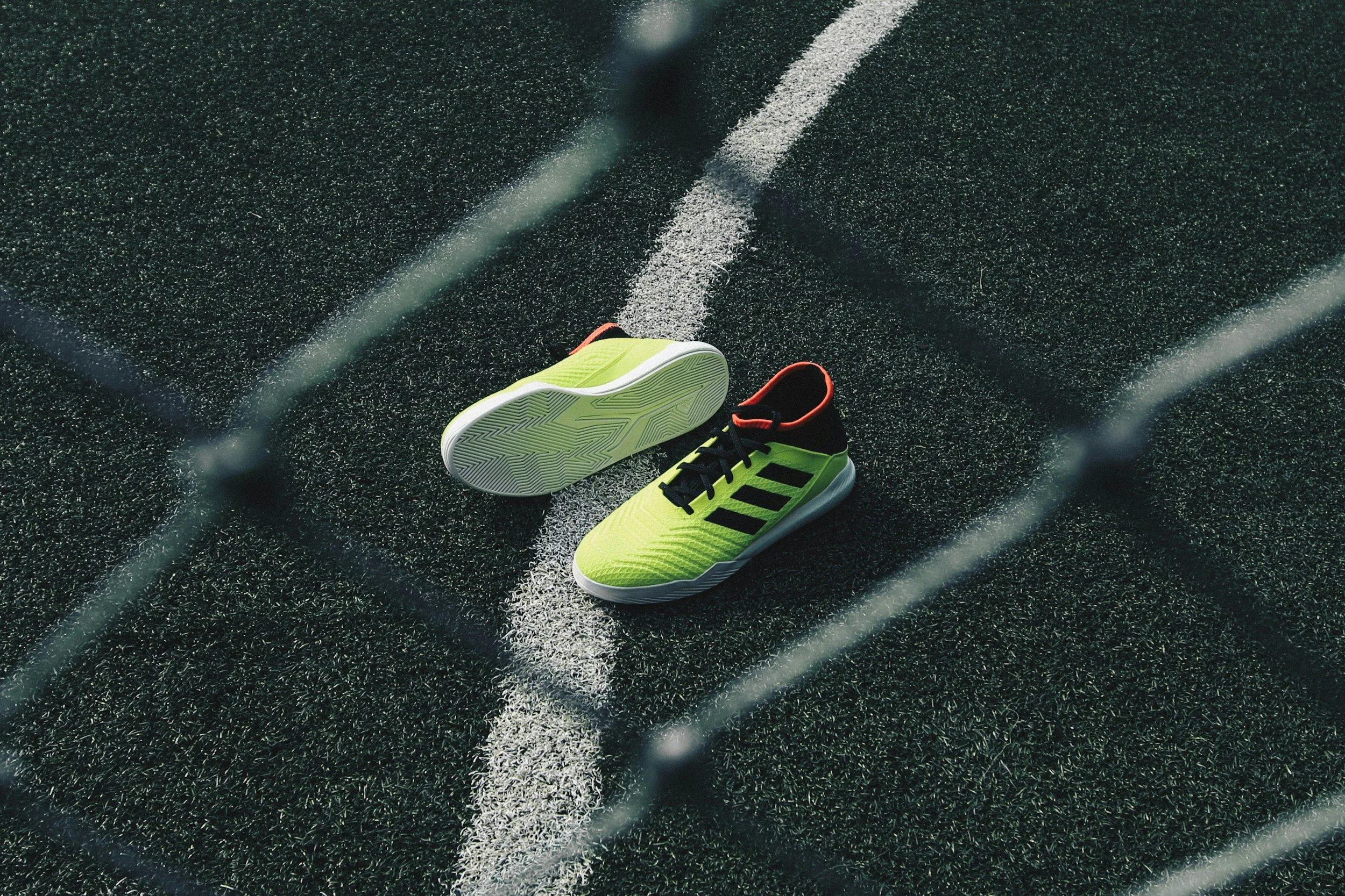Grüne Sportschuhe mit schwarzen Streifen auf einem Fußballplatz, umgeben von einem Maschendrahtzaun.