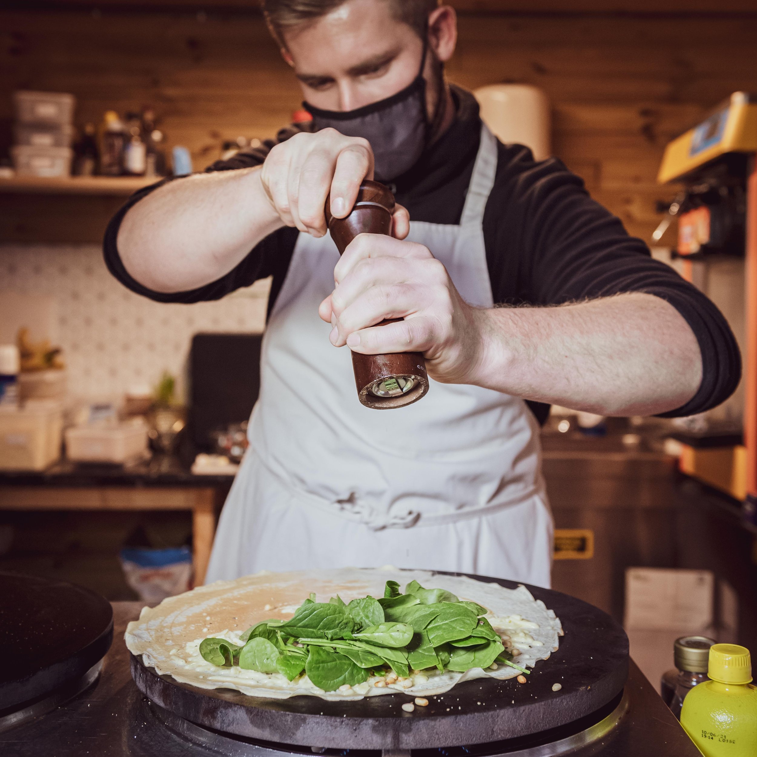 Person seasoning a crepe with spinach on a hot plate in a kitchen, wearing a face mask and apron.