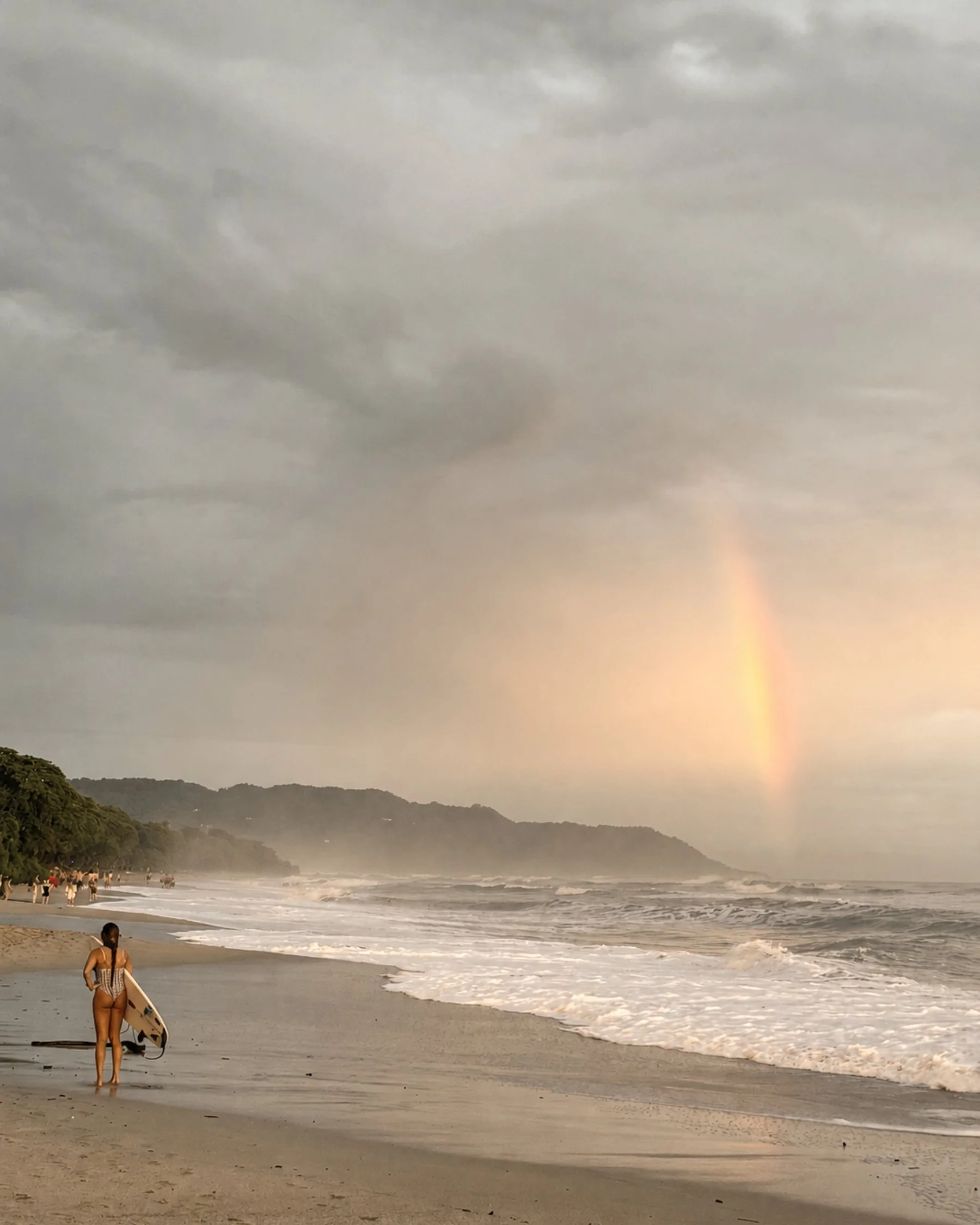 Surfer standing on Santa Teresa beach beneath a rainbow over the Pacific Ocean.