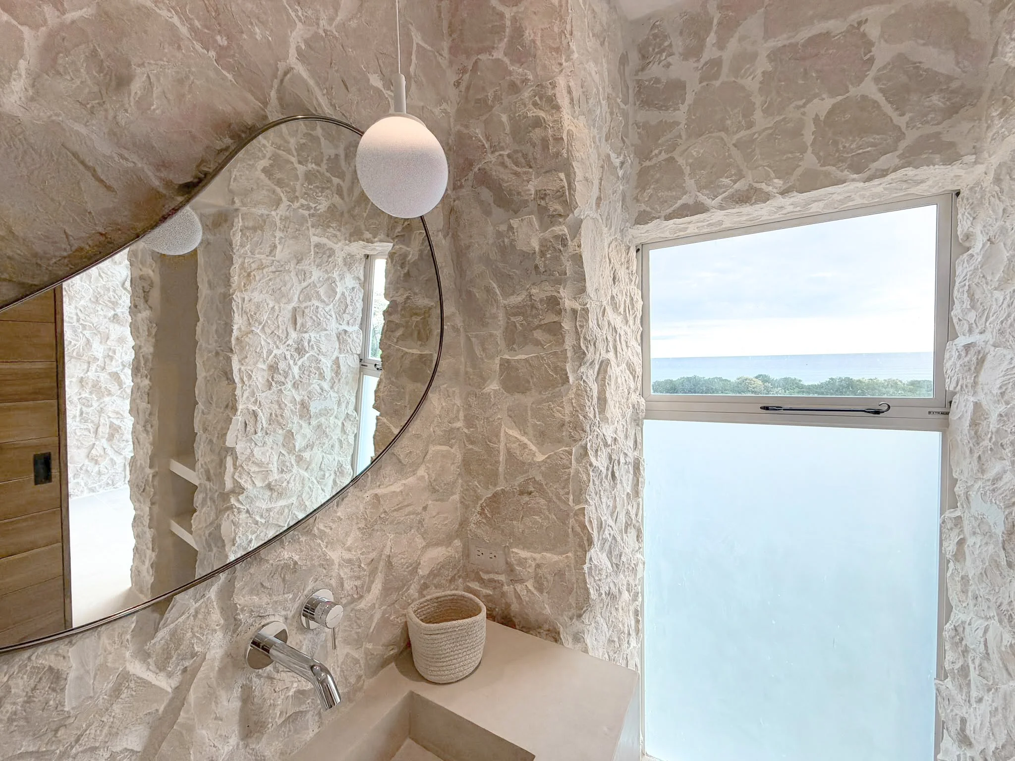 Ensuite bathroom at Casa Cueva with a stone vanity, curved mirror and ocean view through the upper window.