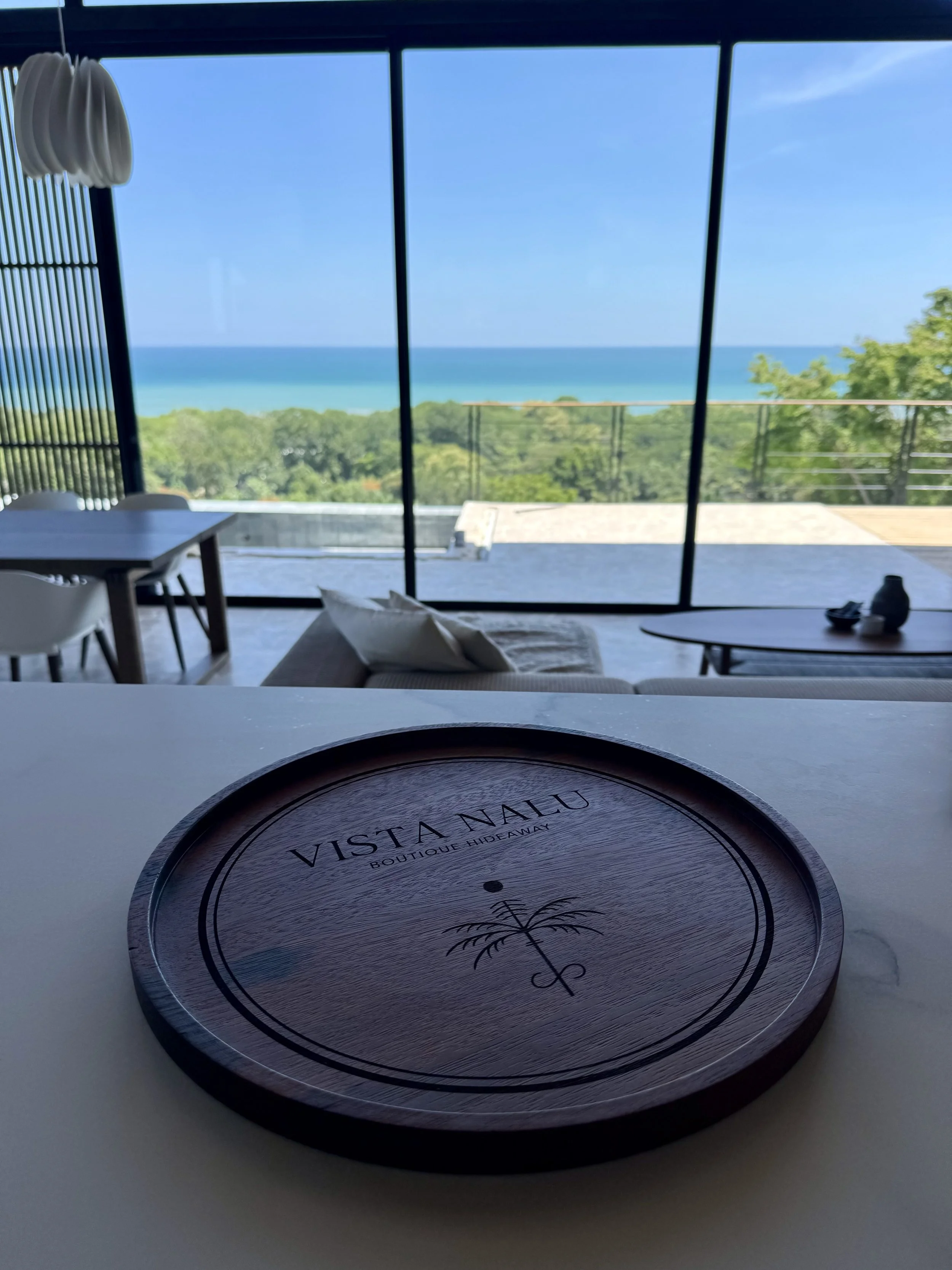 A view from inside a modern cafe or restaurant looking out large glass windows at a scenic ocean view with trees, blue sky, and calm water. In the foreground, a round wooden tray with 'VISTA NALU BOUTIQUE HIDEAWAY' printed on it is on the table, with a beige pillow beside it and a small black object nearby.