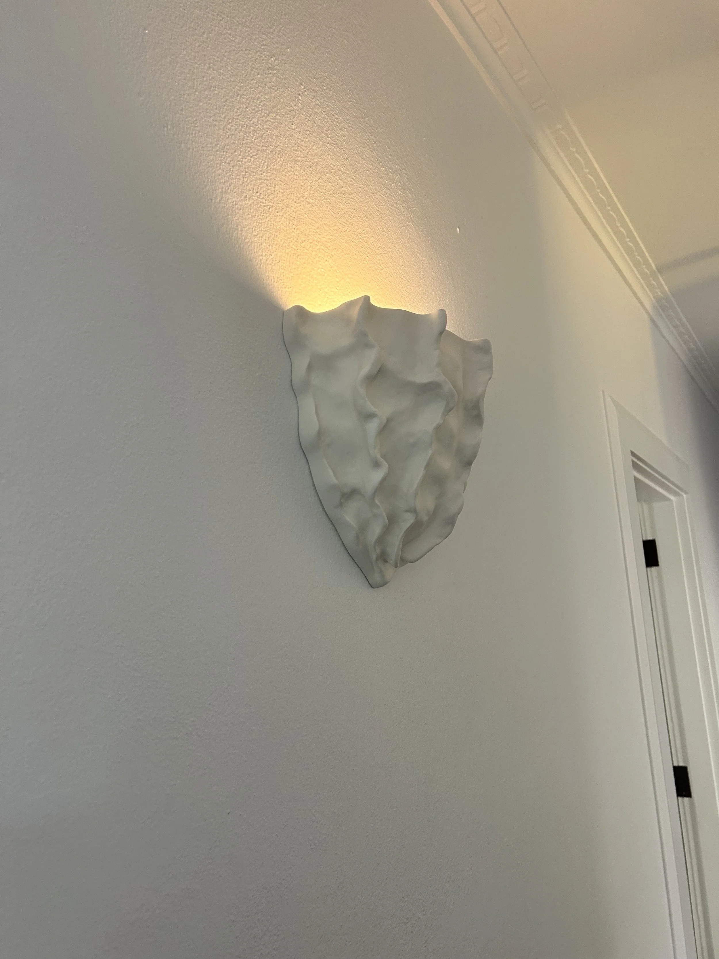 A wall-mounted, abstract white ceramic or plaster sculpture that resembles layered, wavy fabric or organic forms, illuminated from behind.