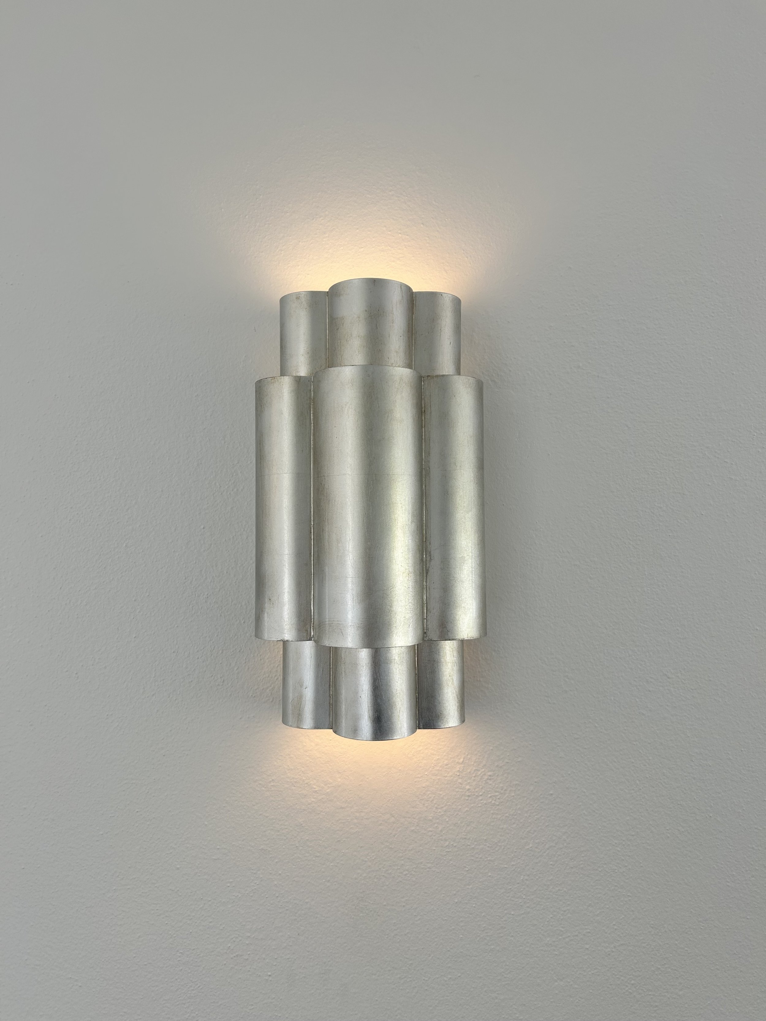 A modern wall sconce with a metallic finish, mounted on a white textured wall, emitting soft light upwards and downwards.