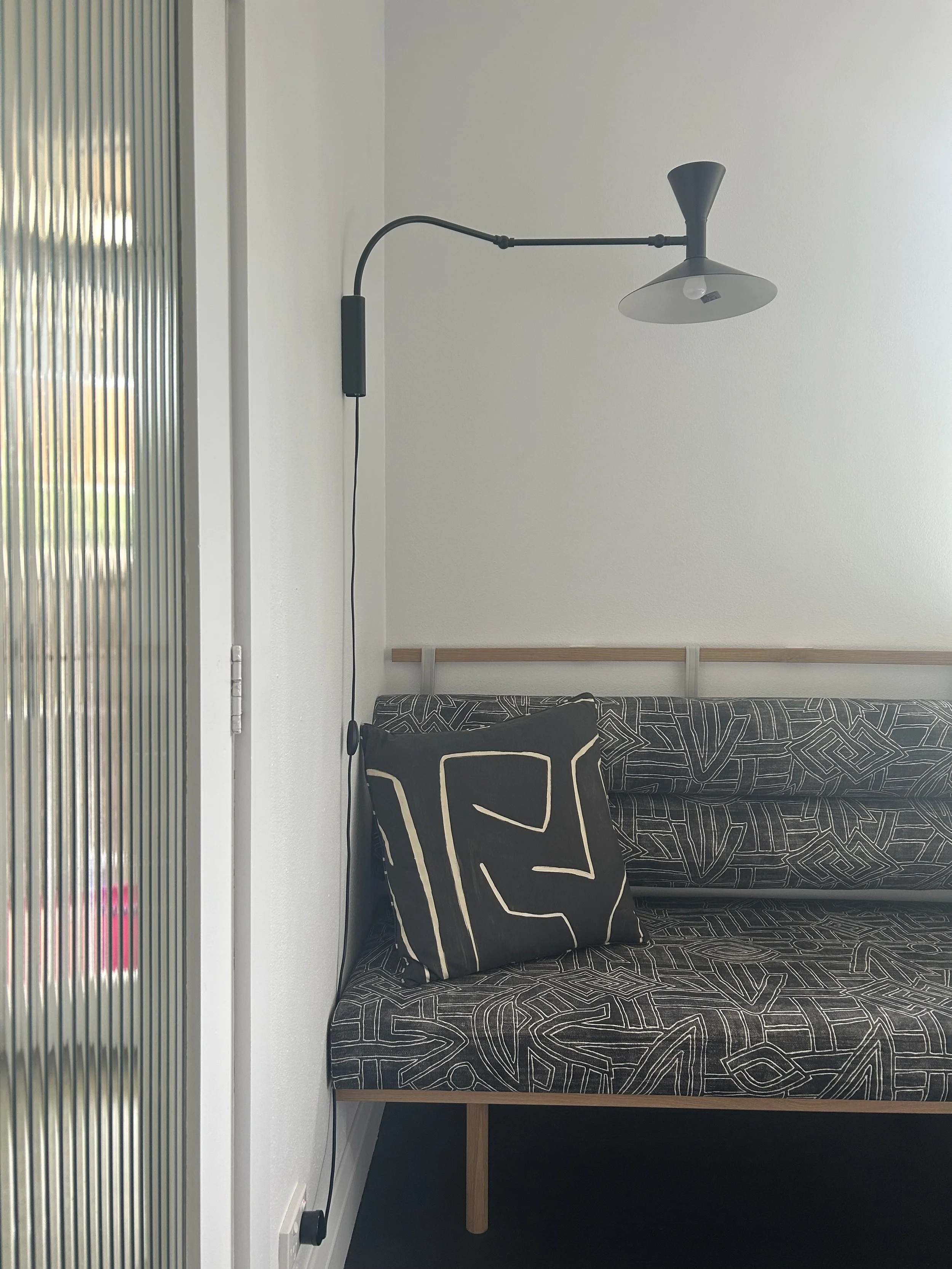 A black wall-mounted lamp above a patterned black and beige sofa with a black and beige pillow in a modern room with a white wall.