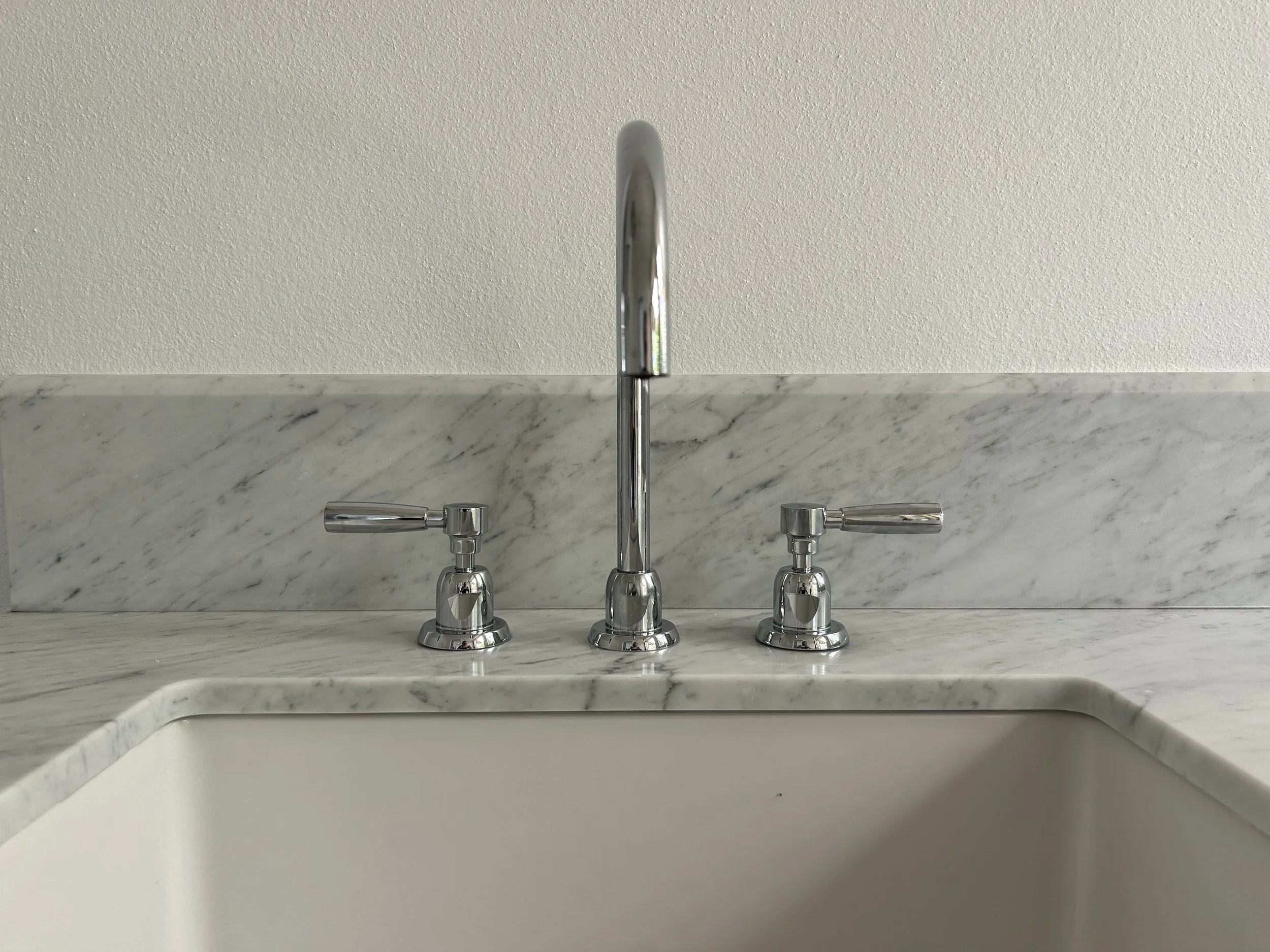 Kitchen sink with a marble countertop, silver faucet, and handles, with a textured white wall in the background.