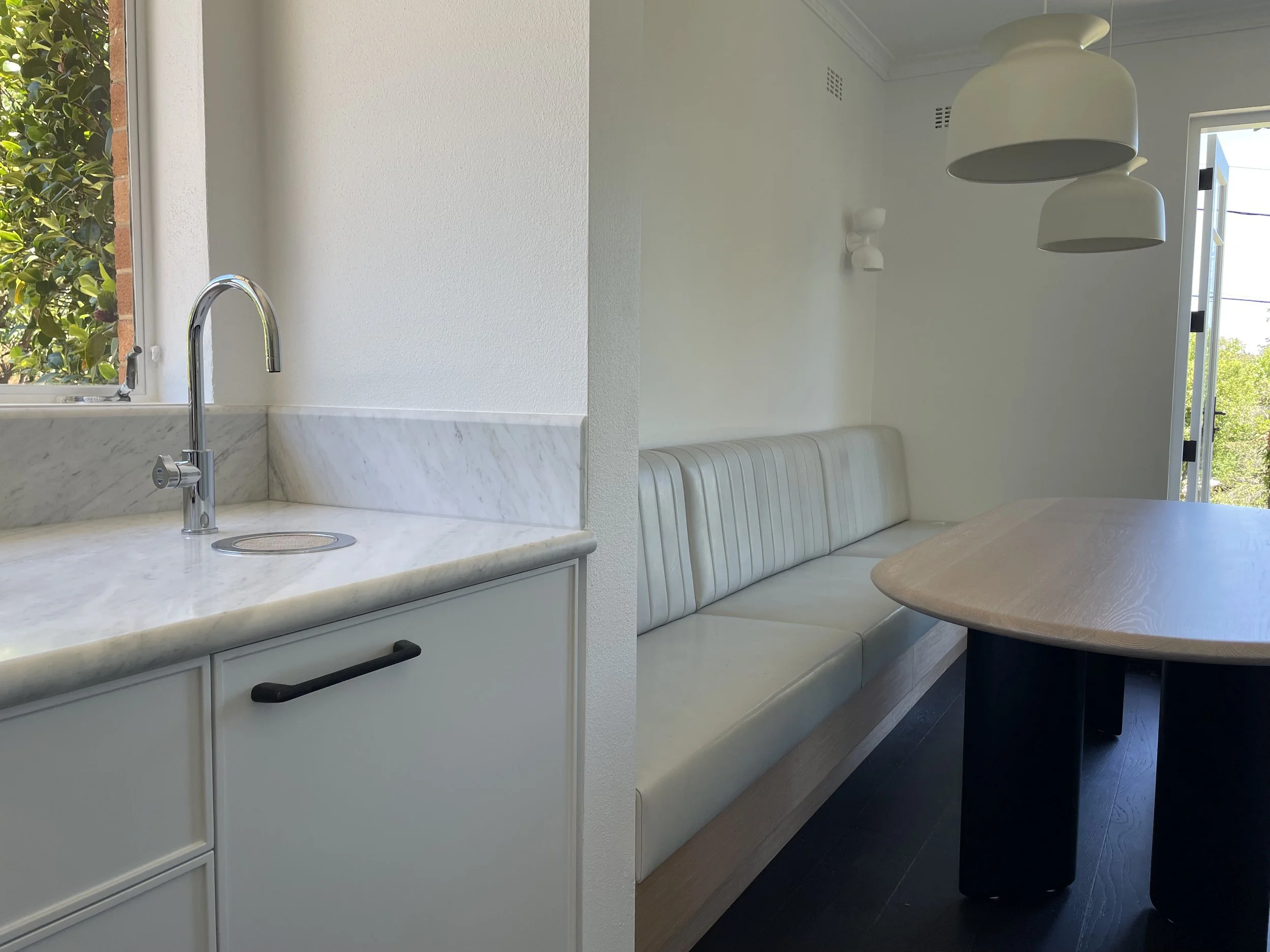 Split image showing a kitchen sink with a window on the left side and a dining nook with a white cushioned bench, a wooden table, and hanging ceiling lights on the right side.