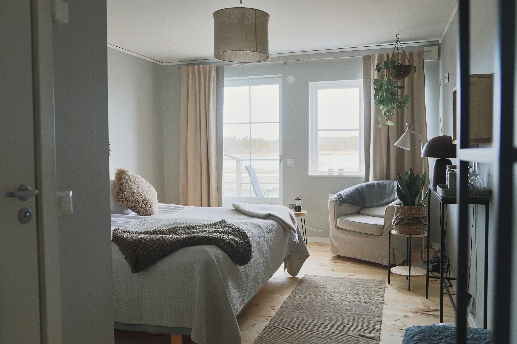 Mysigt sovrum med säng, fåtölj och växter nära ett fönster med gardiner. En neutral färgpalett används genomgående, med en beige matta, filtar och en hängande lampa.