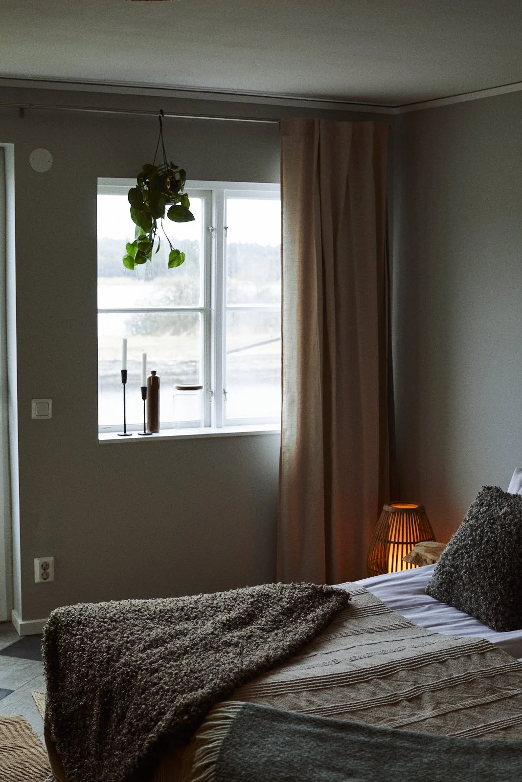 Mysigt sovrum med ett fönster, en hängande växt, en säng med filtar och en flätad lampa.