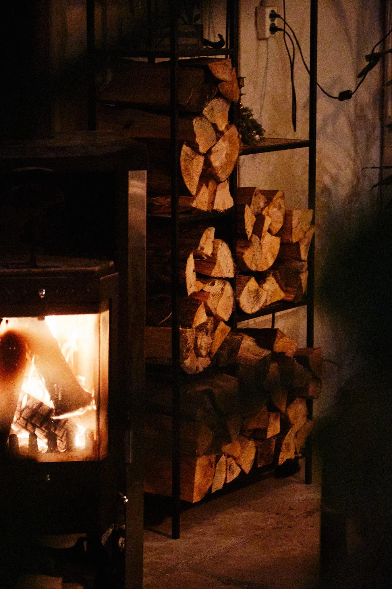 Cozy indoor scene with a lit wood stove and stacked firewood on shelves.