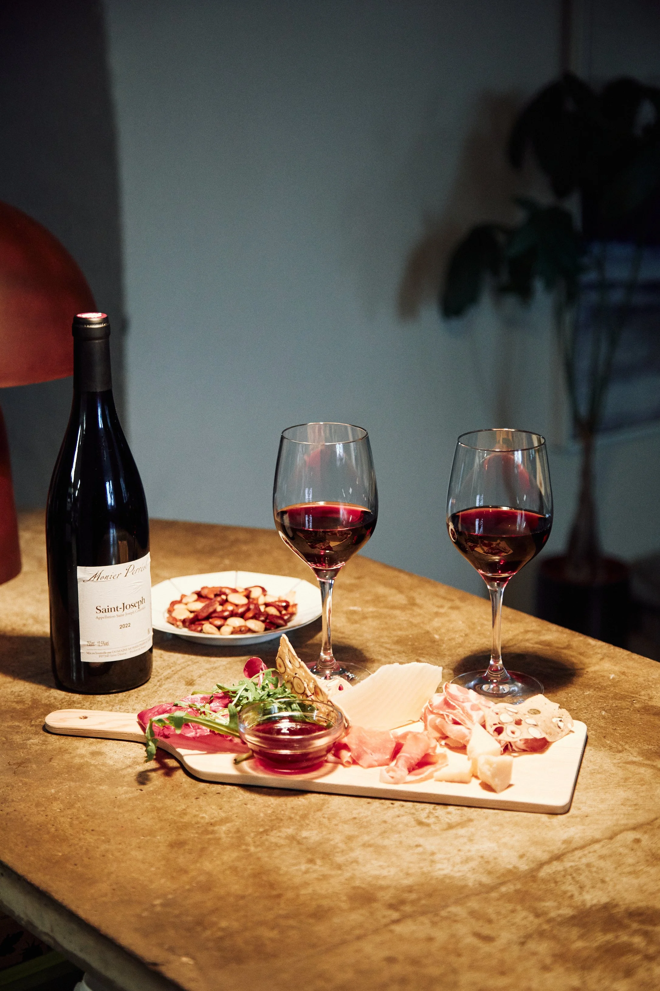 Two wine glasses filled with red wine, a wine bottle labeled "Saint-Joseph 2022," and a wooden charcuterie board with assorted meats, cheese, and greens, on a table; a bowl of sliced nuts in the background.