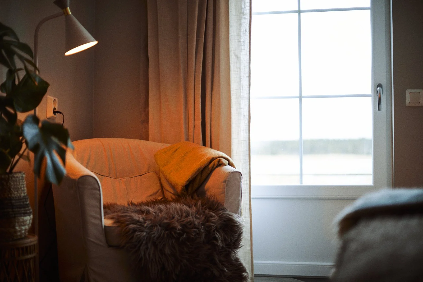Mysig vardagsrumshörna med en fåtölj, en lampa, ett fönster med gardiner och en planta.