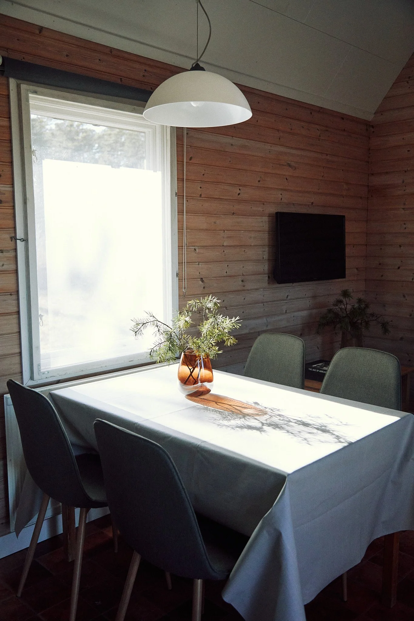 Dining room with a wooden wall, white tablecloth, vase with green branches, pendant lamp, and gray chairs. middagsbord I stugorna som är perfekt för hela familjen.