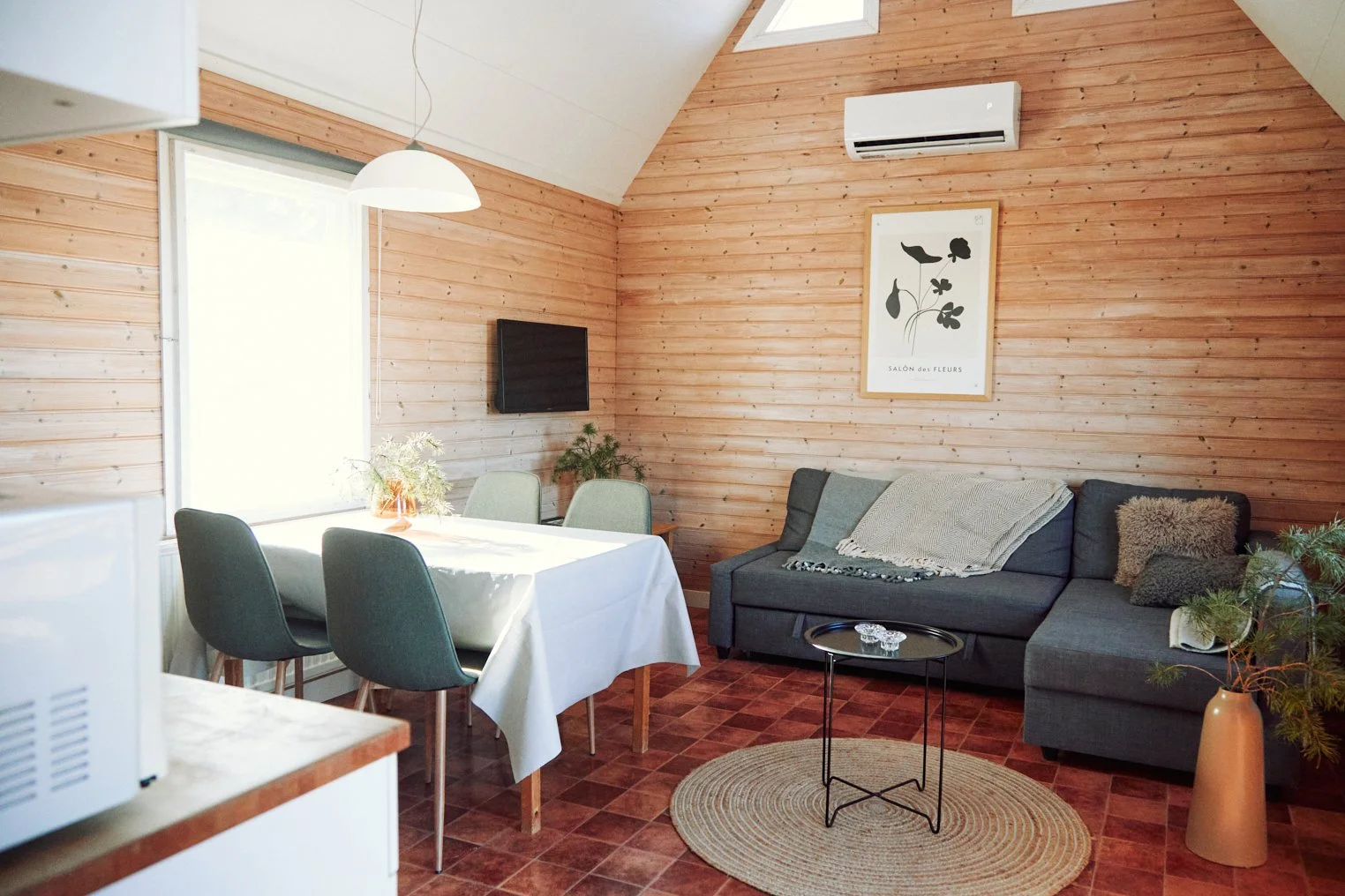 Mysigt vardagsrum med träväggar, en grå soffa, litet svart soffbord och en matplats med gröna stolar och ett dukklätt bord. Minimalistisk inredning med växter och väggkonst. vardagsrum