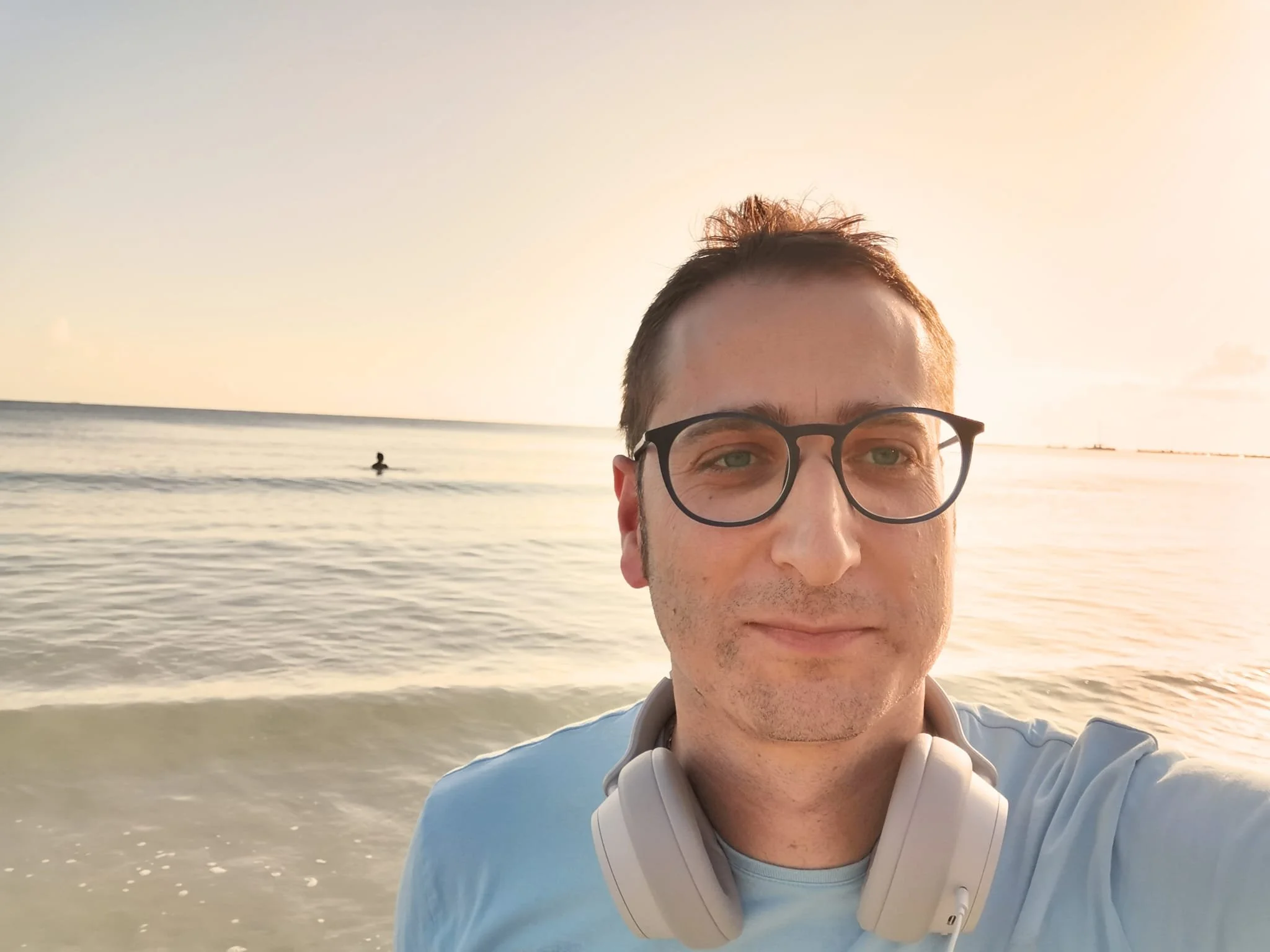 A man wearing glasses and headphones taking a selfie at the beach during sunset, with a person swimming in the ocean in the background.