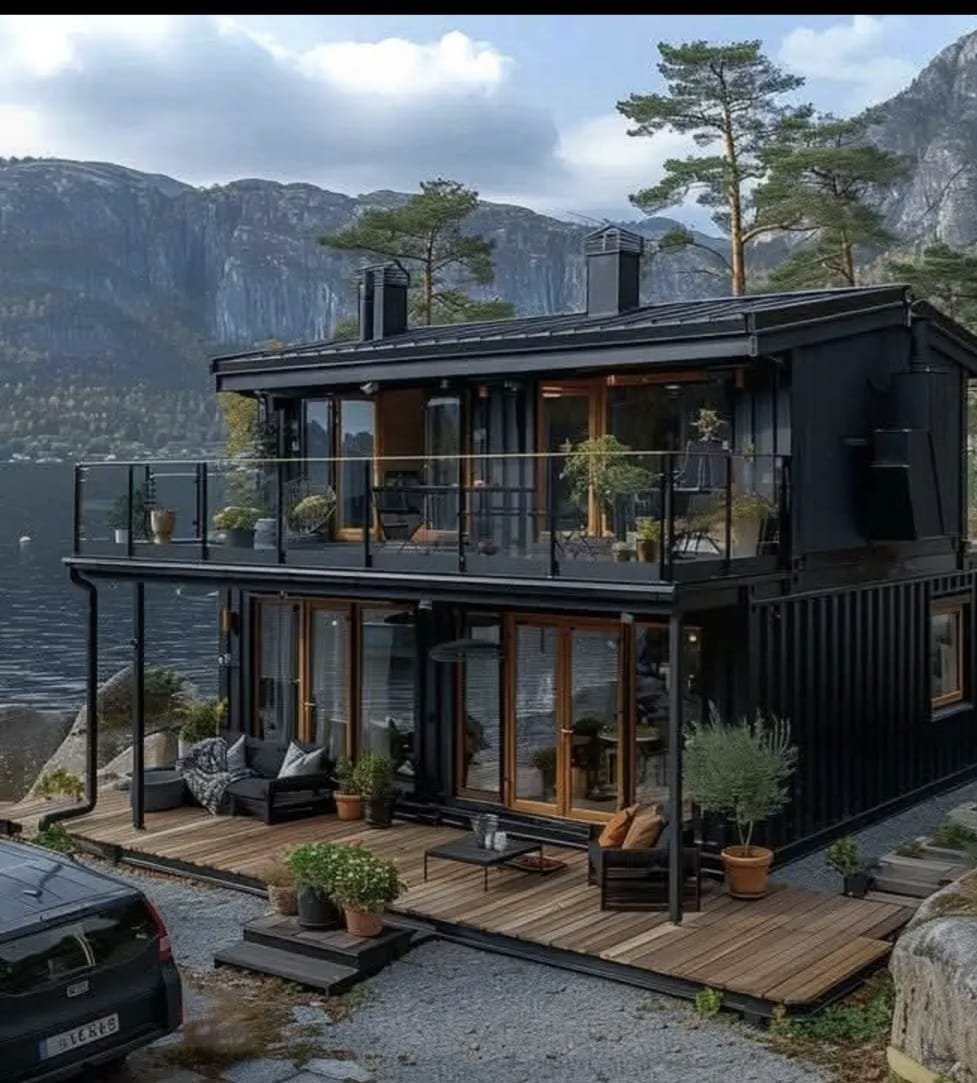 Modern black two-story modular container house with large glass windows and balconies, situated near a lake with mountains in the background, surrounded by trees and outdoor furniture on wooden decks.