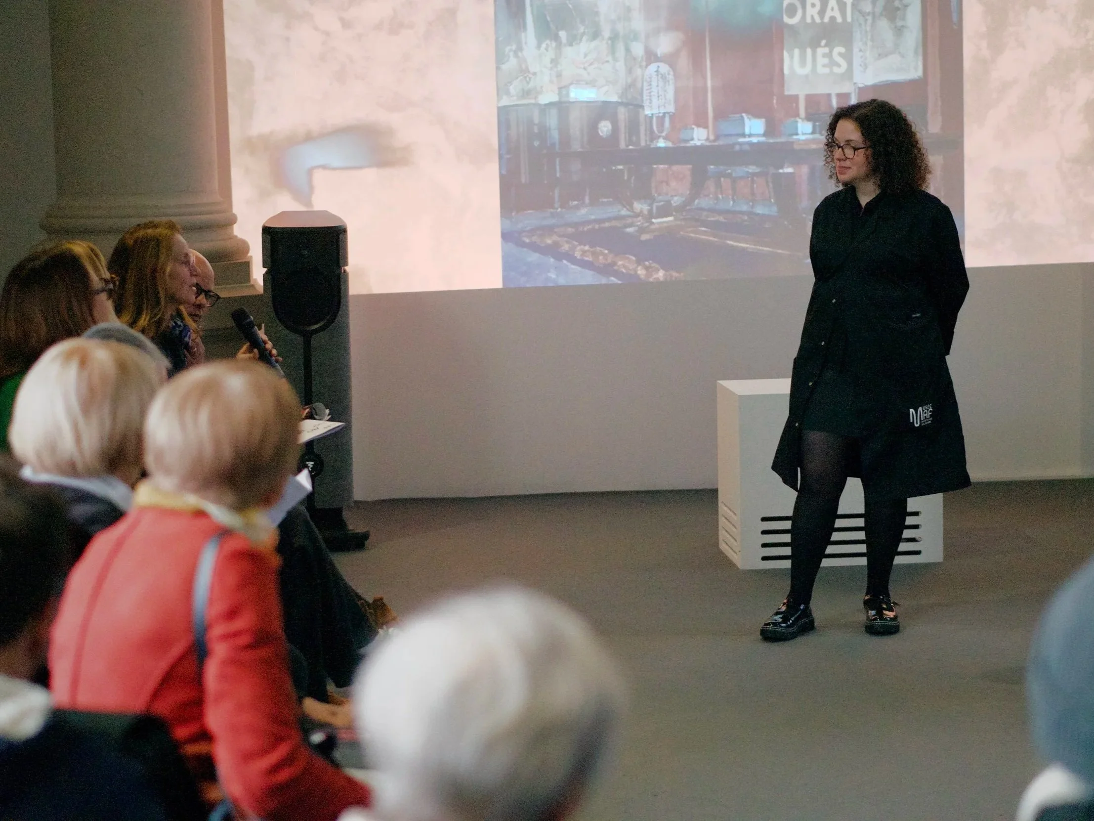 Une femme avec des cheveux bouclés noirs et des lunettes, portant une robe noire, se tient devant un public lors d'une présentation ou conférence, avec un écran derrière elle affichant une image d'une pièce avec un piano et une bannière.