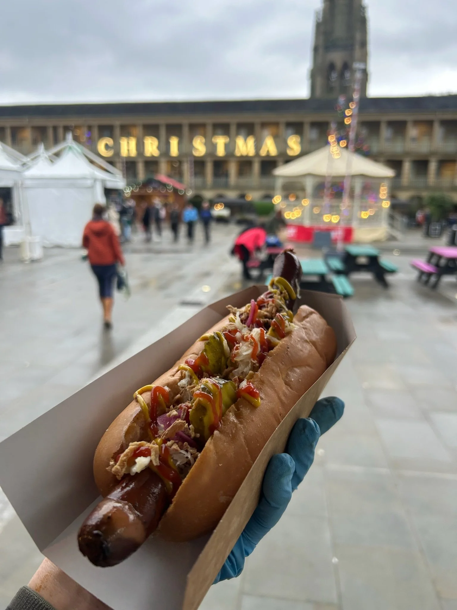 Hot dog with toppings in front of a Christmas market, with a building and Christmas lights in the background.