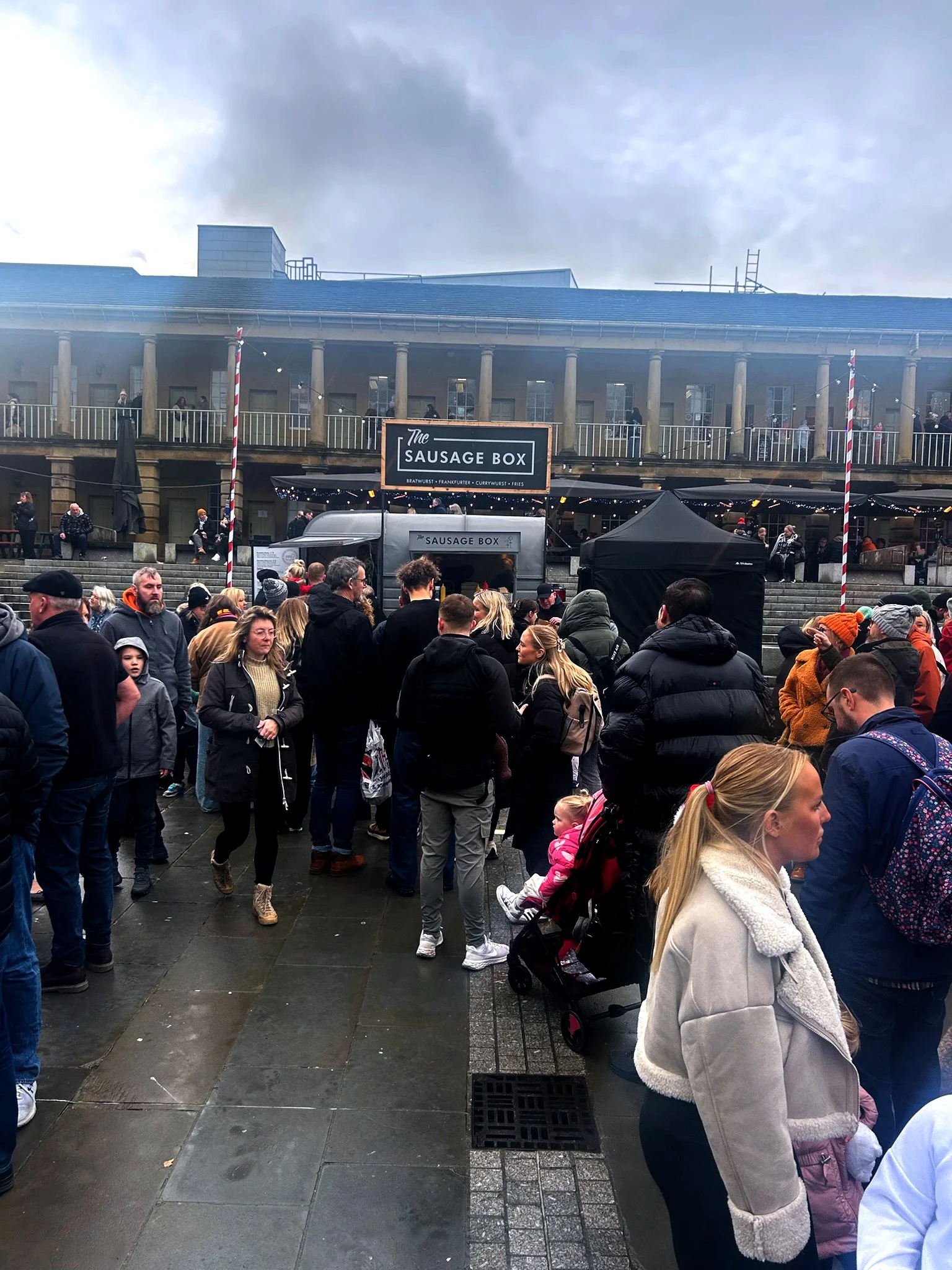 Crowd of people waiting in line at The Sausage Box food stand outdoors, with a building and cloudy sky in the background.
