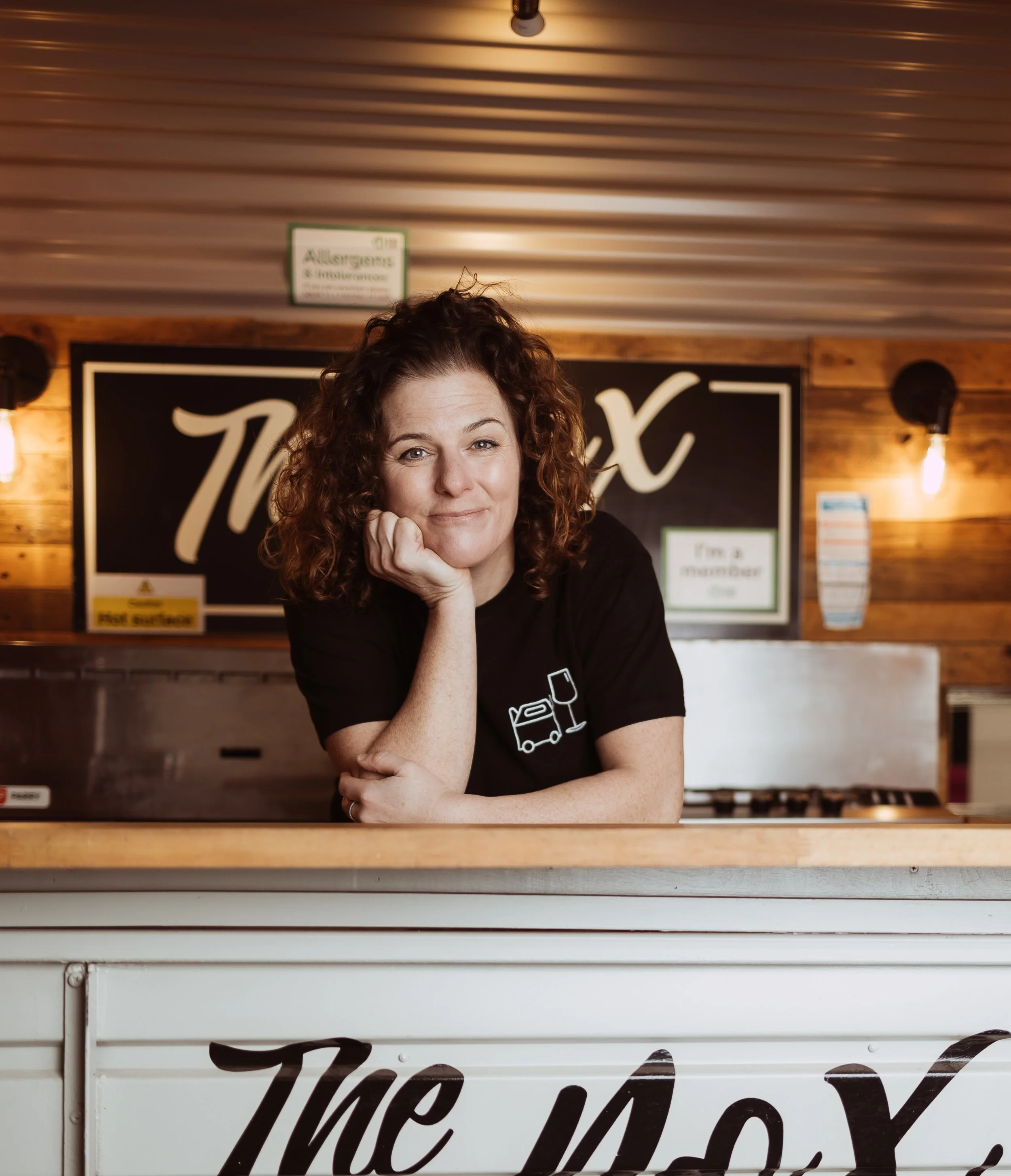 A woman with curly hair sitting behind a counter in a restaurant or bar, with a sign in the background and wooden wall paneling.