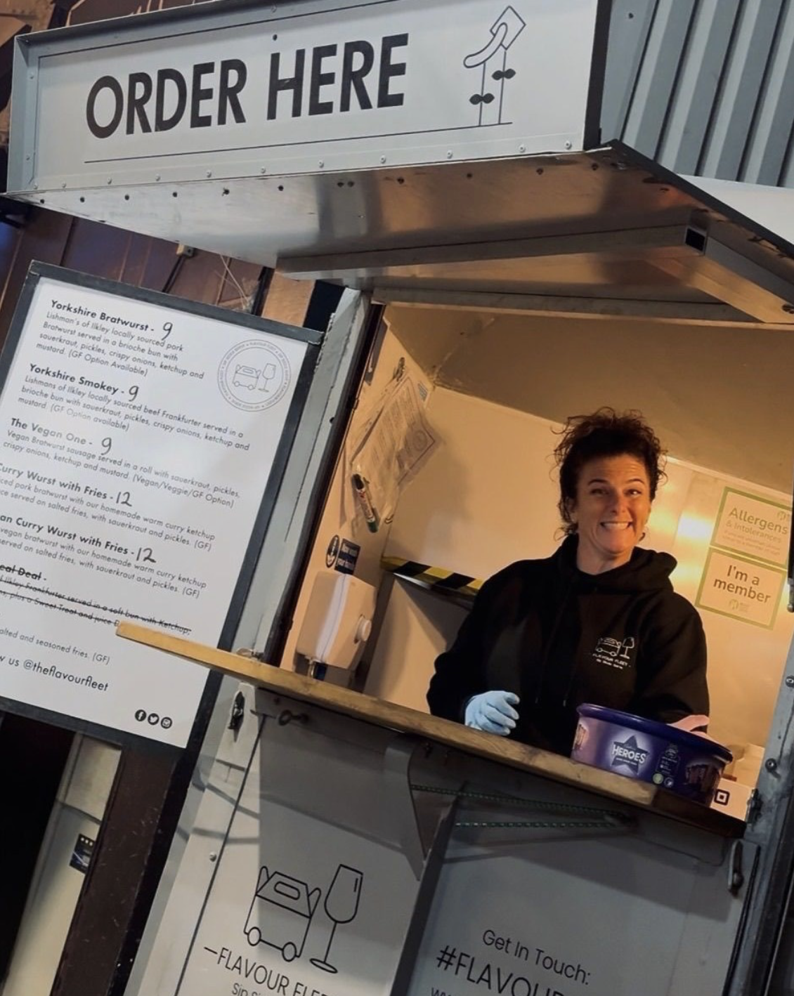 A woman at a food stand smiling with a black hoodie and gloves, behind a counter with a menu listing sausage options, inside a small food truck with signs on the wall.