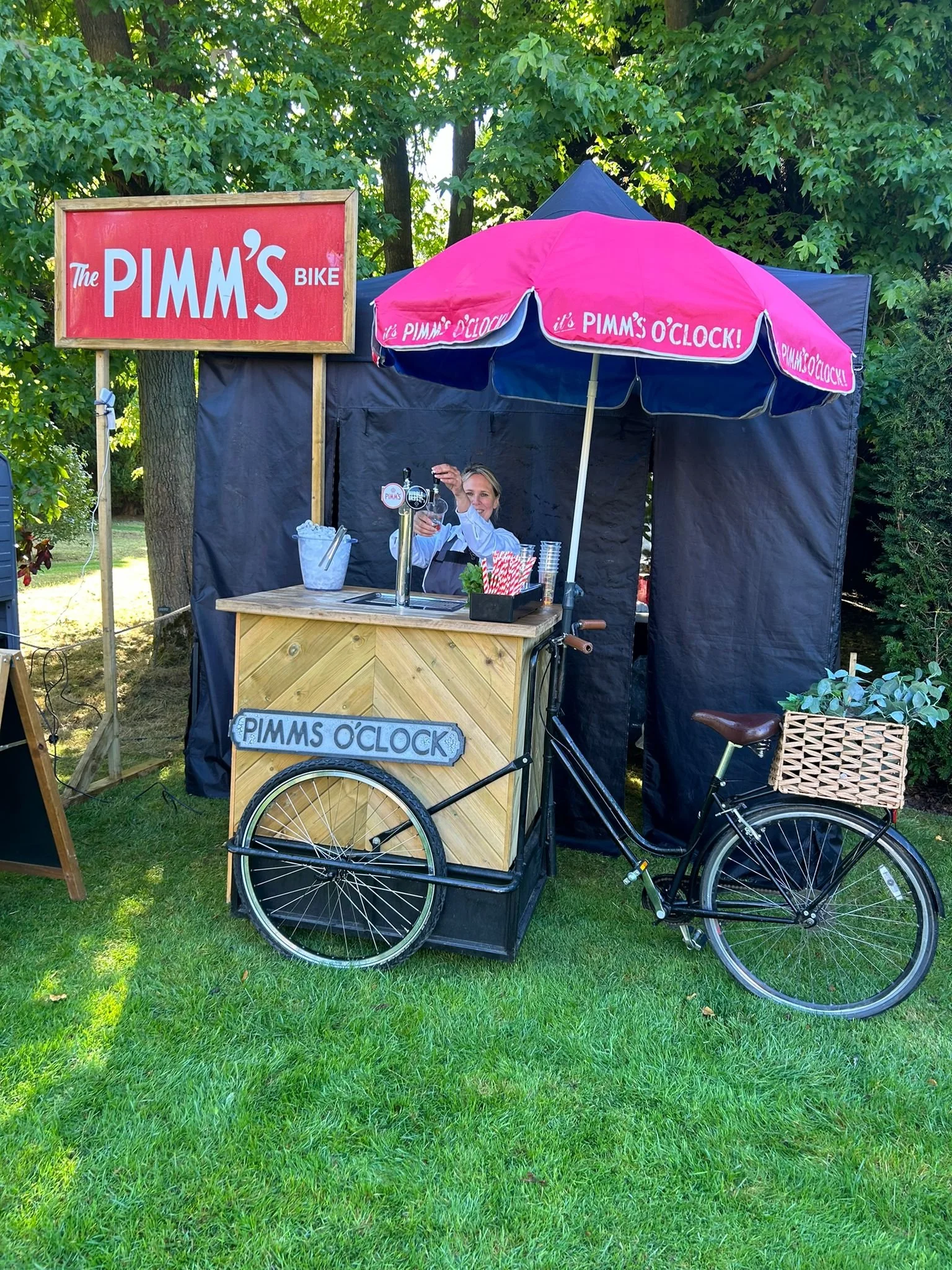 A woman behind a small outdoor bar stall called 'Pimm's O'Clock' with a pink and blue umbrella, a bicycle with a basket, and a sign that reads 'PIMM'S BIKE' in a green park setting.