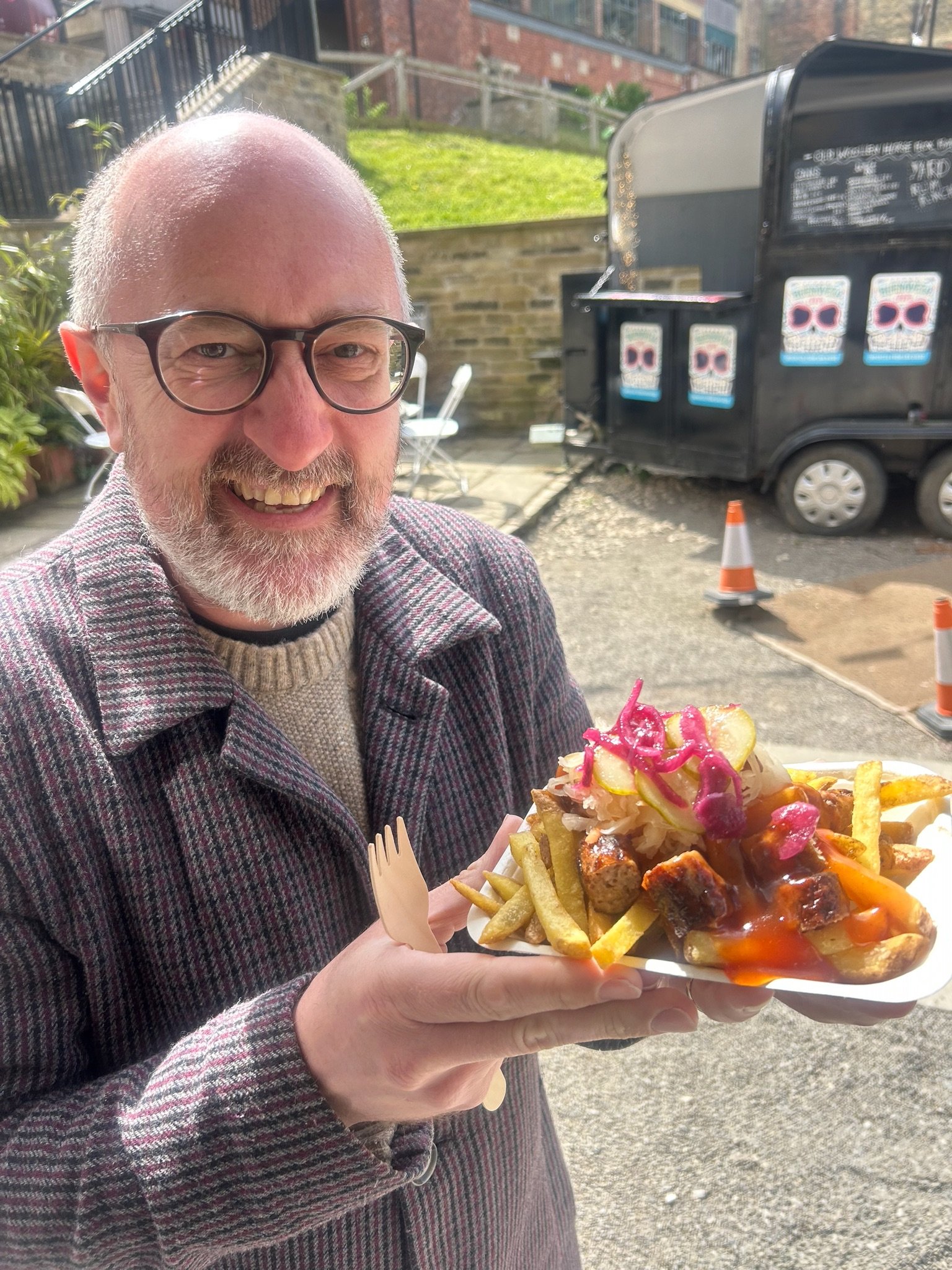 A smiling man with glasses holding a tray of loaded fries topped with pickles, pink cabbage, sauce, and meat. He is outdoors with a food truck and traffic cones in the background.