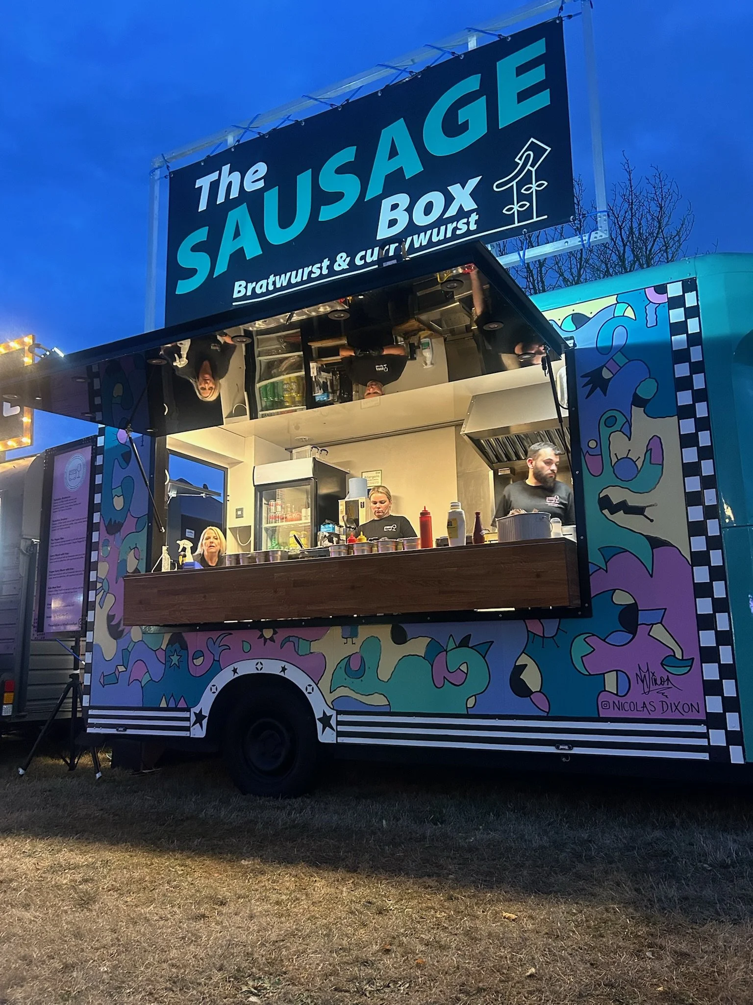 A food truck with a colorful, abstract design on its exterior, serving brats, wursts, and currywurst in an outdoor setting during early evening. The truck has a blue and purple mural with various shapes and features a sign reading "The Sausage Box."