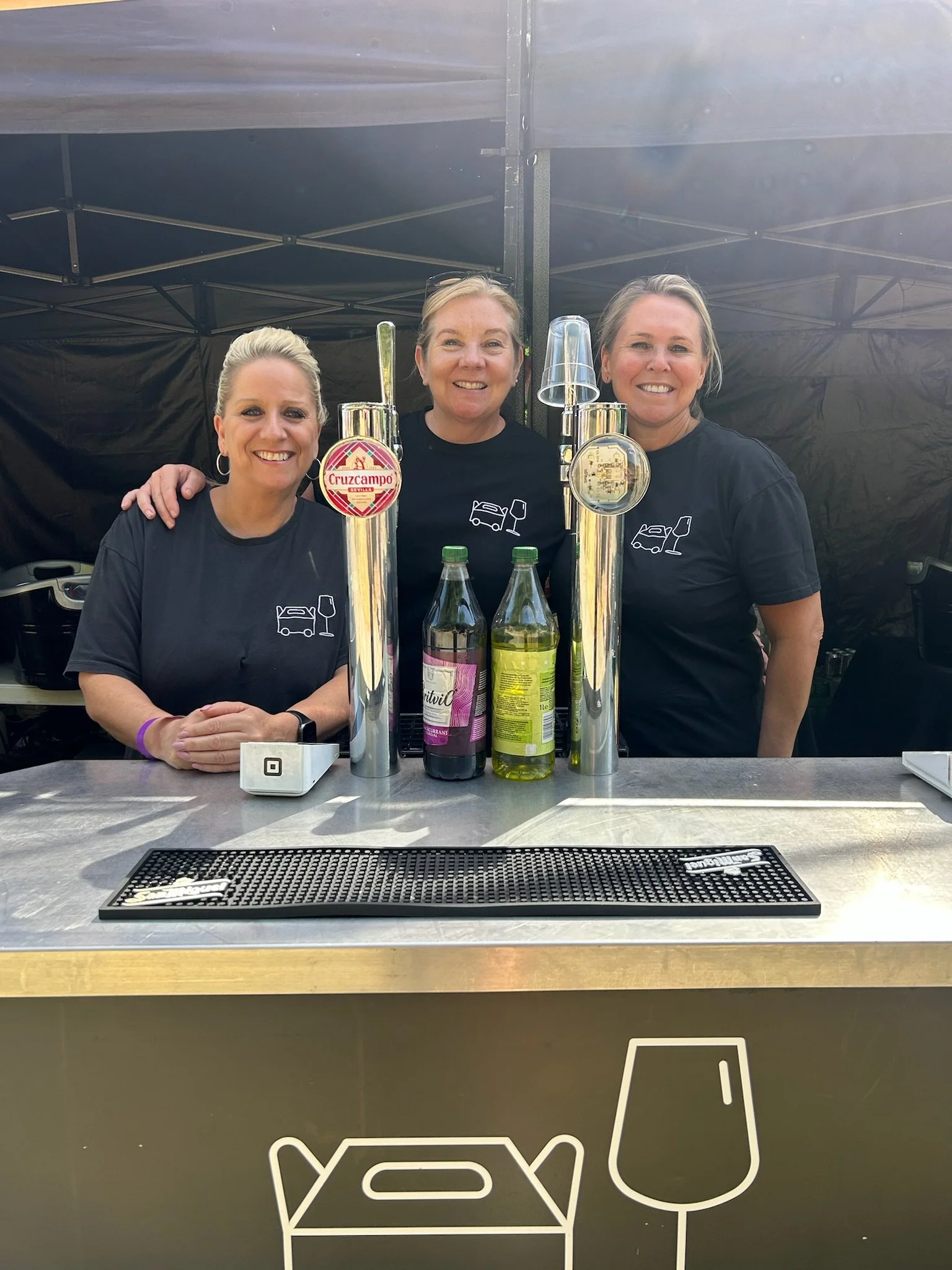 Three women standing behind a bar counter with beer taps, bottles, and glasses, under a black canopy.