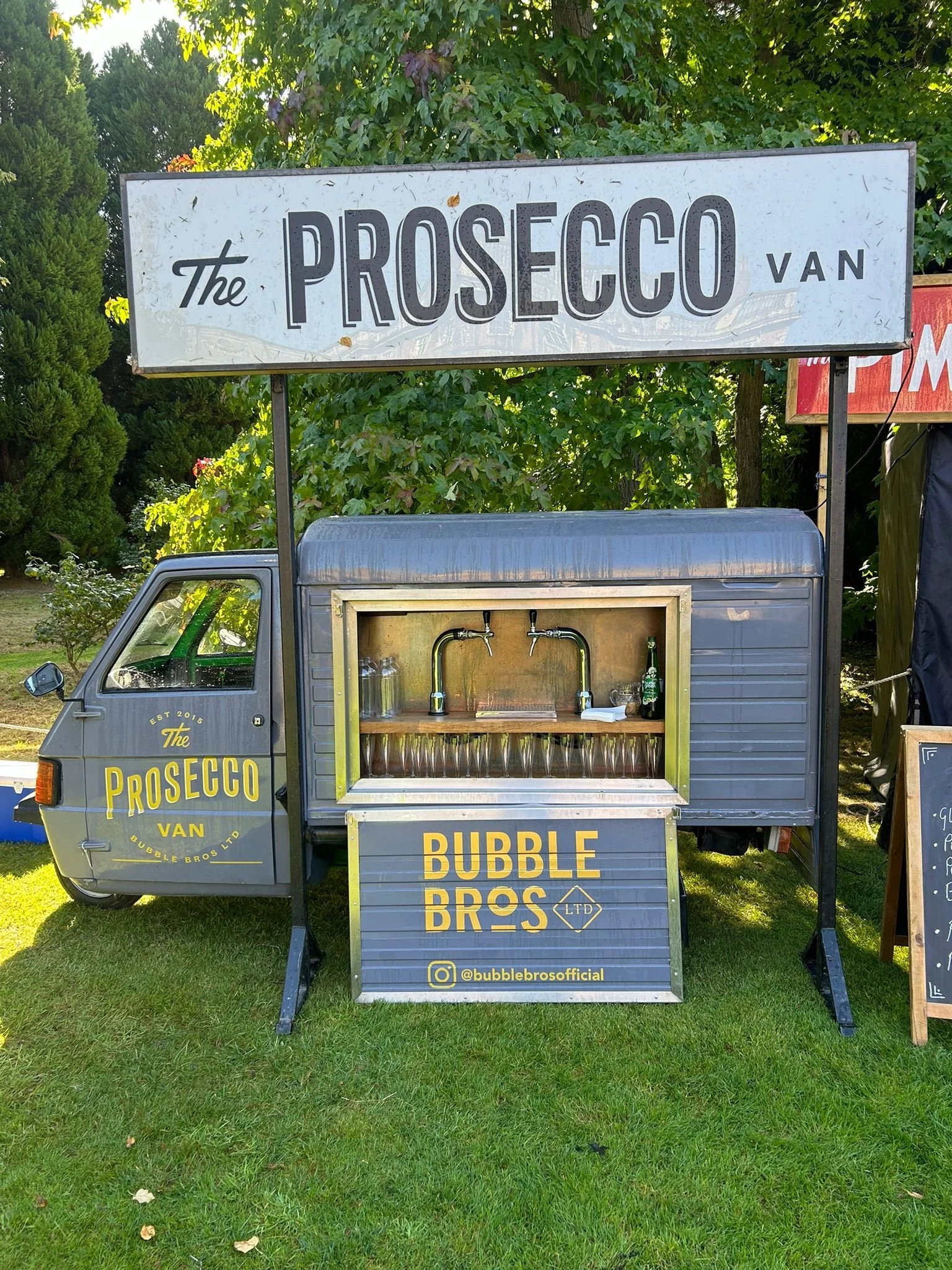 A small gray Prosecco van stand with a serving window for glasses and a tap, branded with yellow 'Bubble Bros' logo and Instagram handle, set on green grass with trees in background.