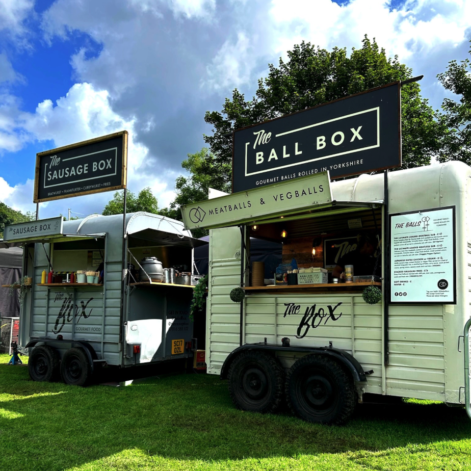 Two food trucks named The Sausage Box and The Ball Box parked on grass, with trees and a partly cloudy sky in the background.