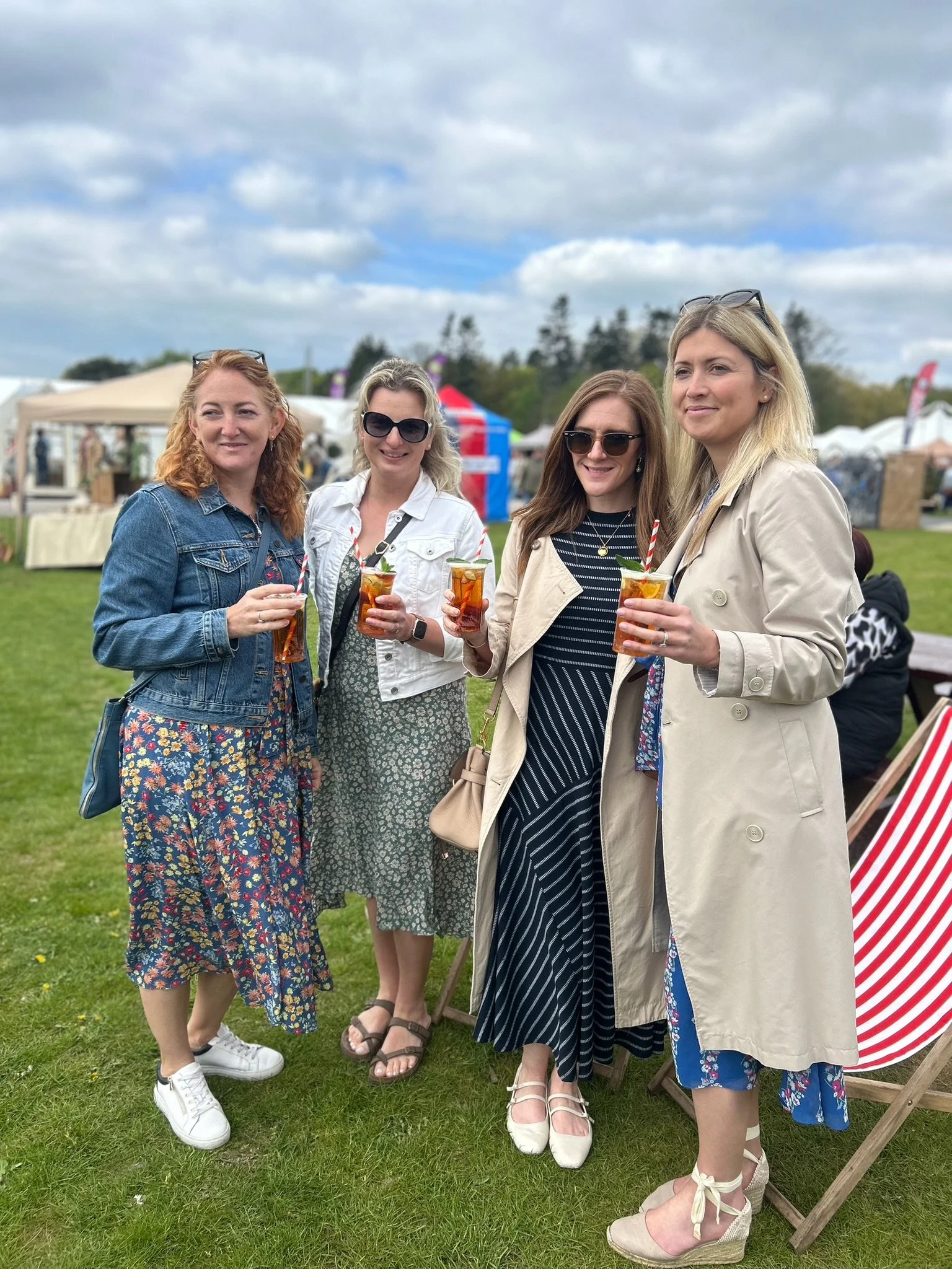 Four women standing together outdoors at a festival, holding drinks, smiling, with tents and a cloudy sky in the background.
