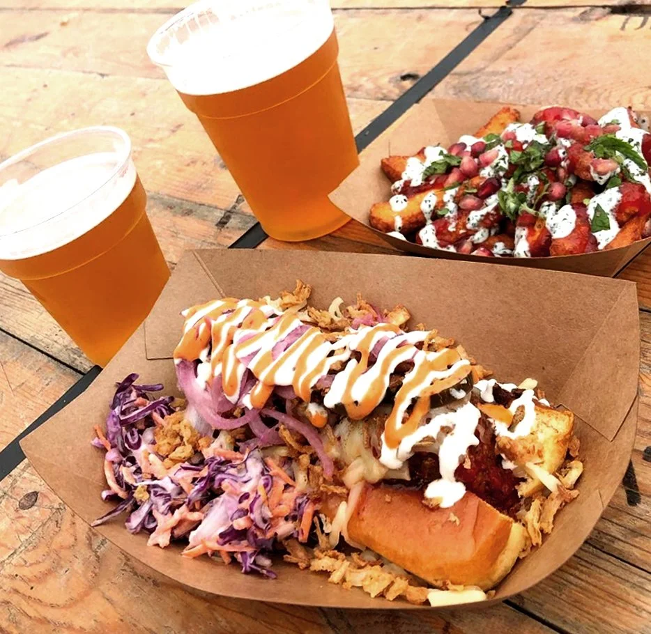 Two cups of beer, a plate with a loaded hot dog topped with sauces and onions, and another plate with loaded fries topped with cheese, herbs, and sauces on a wooden table.