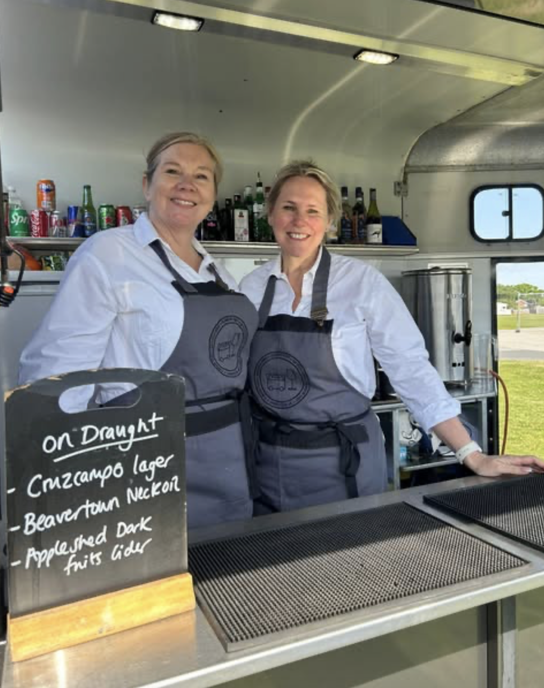 Two women standing behind a bar counter with drinks and a chalkboard sign that reads 'on Draught, Cruzcampo Lager, Beavertown Neck Oil, Applehashed Dark Fruit Beer'. They are smiling and wearing white shirts with gray aprons.