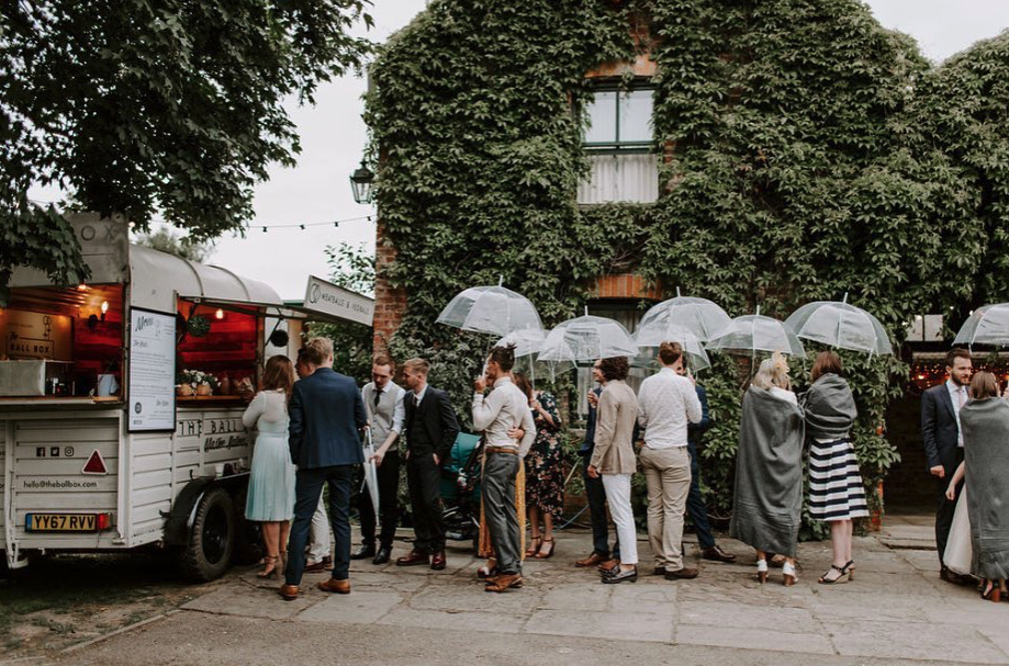 People standing in line with umbrellas outside a food truck on a city street, surrounded by greenery and a brick building.