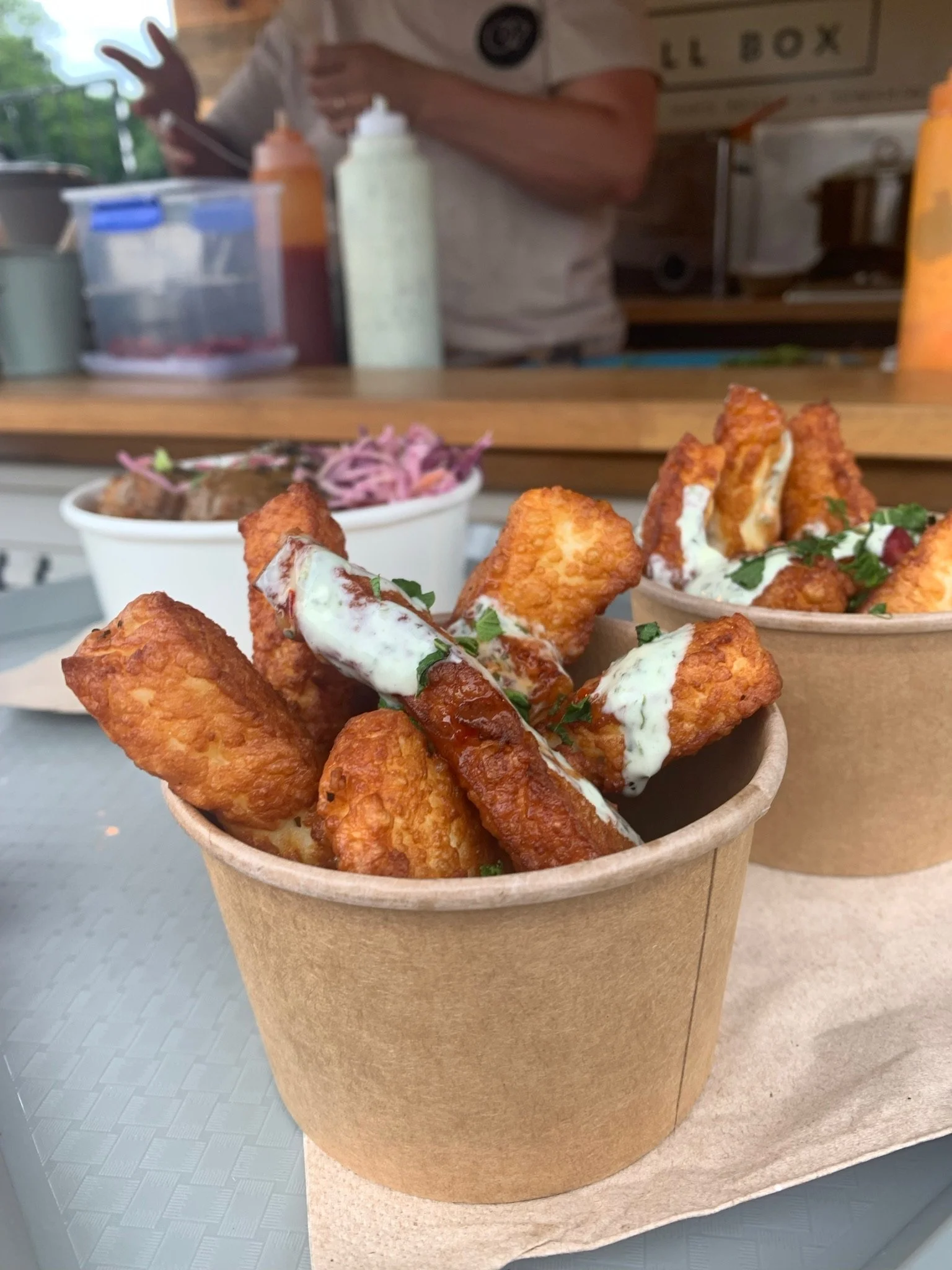 Fried fish sticks with white sauce and herbs in a paper cup, with a side of coleslaw in a separate cup in the background.