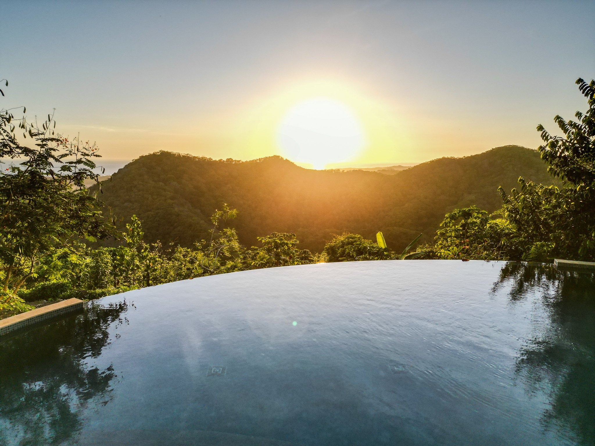 Infinity pool with view of lush green hills and sunrise or sunset sky.