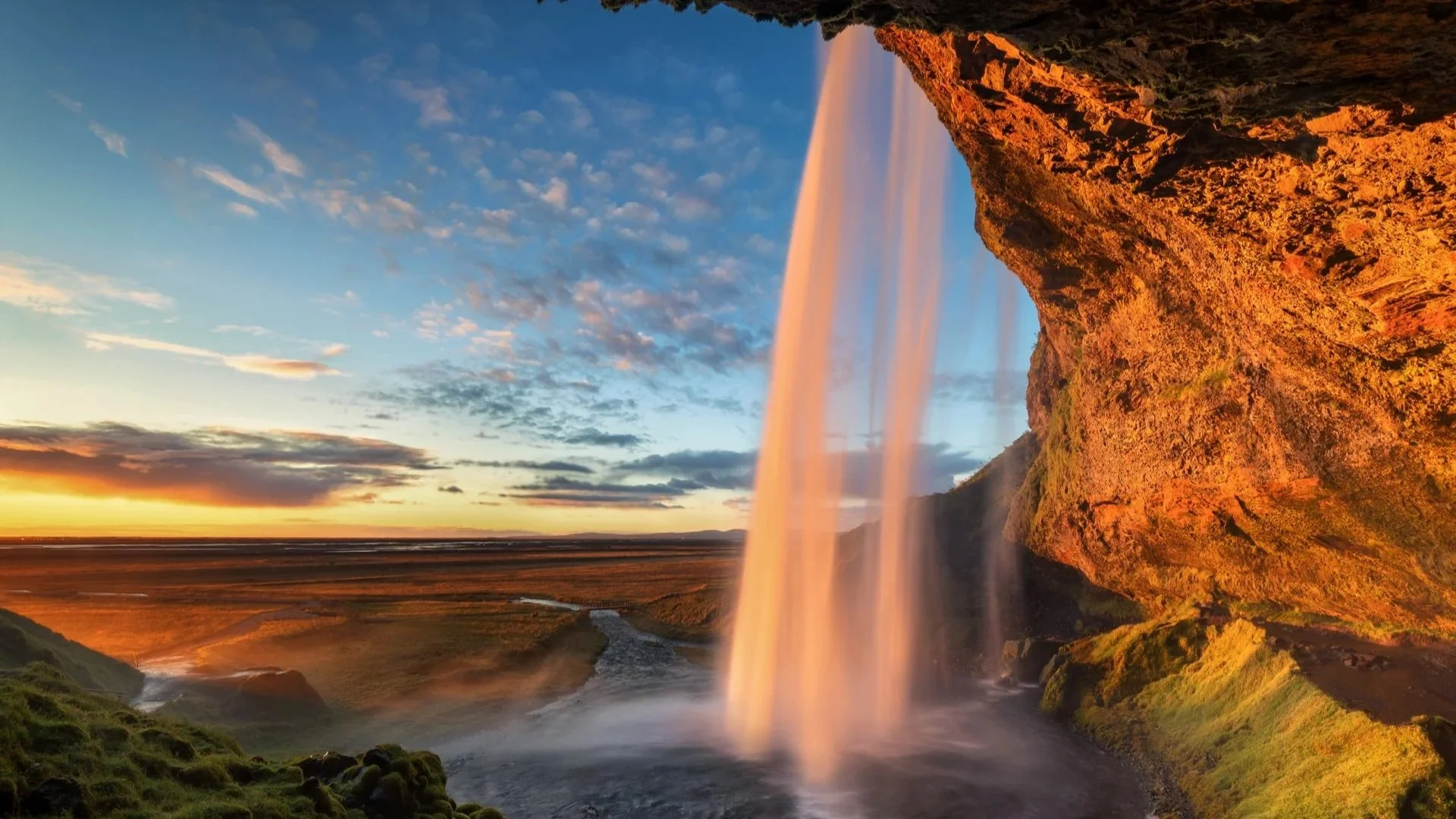 Ein Wasserfall während des Sonnenuntergangs mit rotem Licht, Wasser strömt von einer Klippe in eine Flusslandschaft, im Hintergrund ein Himmel mit Wolken und die Sonne, die den Horizont beleuchtet.