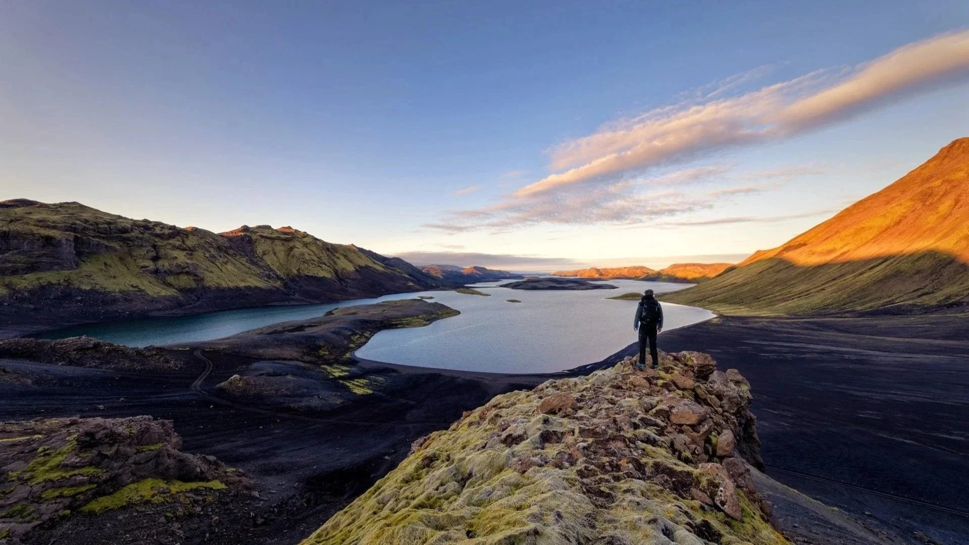 Ein einzelner Wanderer mit Rucksack steht auf lavabedecktem Boden vor einem See, umgeben von grünen und schwarzen Bergen, im Sonnenuntergang bei klarem Himmel.