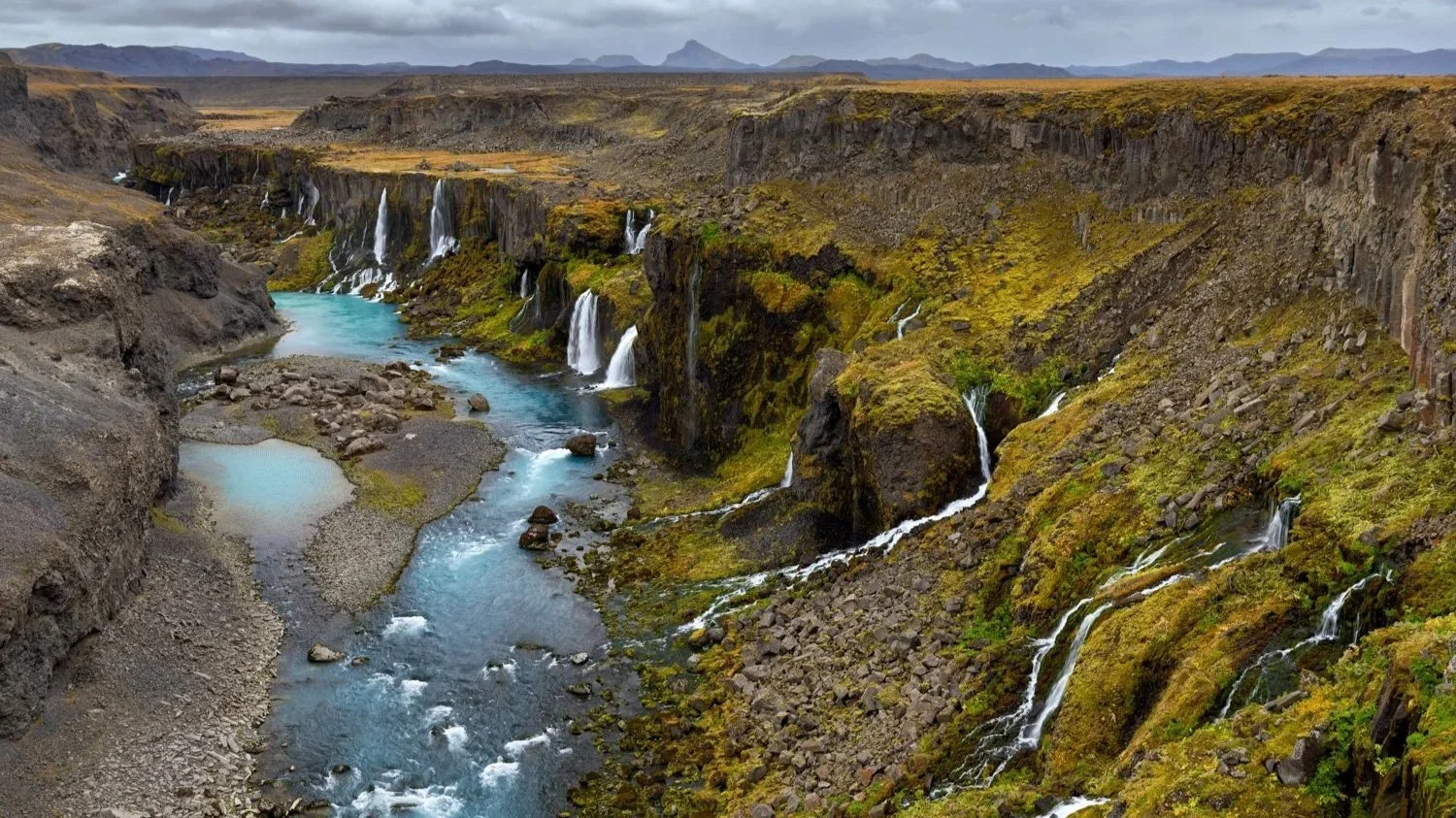 Eine beeindruckende Panoramaaufnahme eines Flusstals mit mehreren Wasserfällen, umgeben von grünen, moosbedeckten Felsen in einer kargen, vulkanischen Landschaft. Der Himmel ist bewölkt.