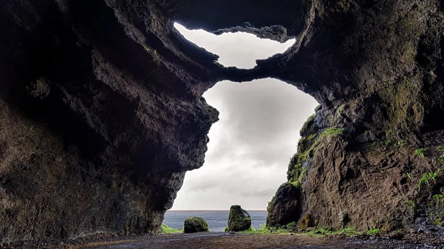 Blick aus einer Höhle auf den Ozean mit Felsen im Wasser und bewölkten Himmel