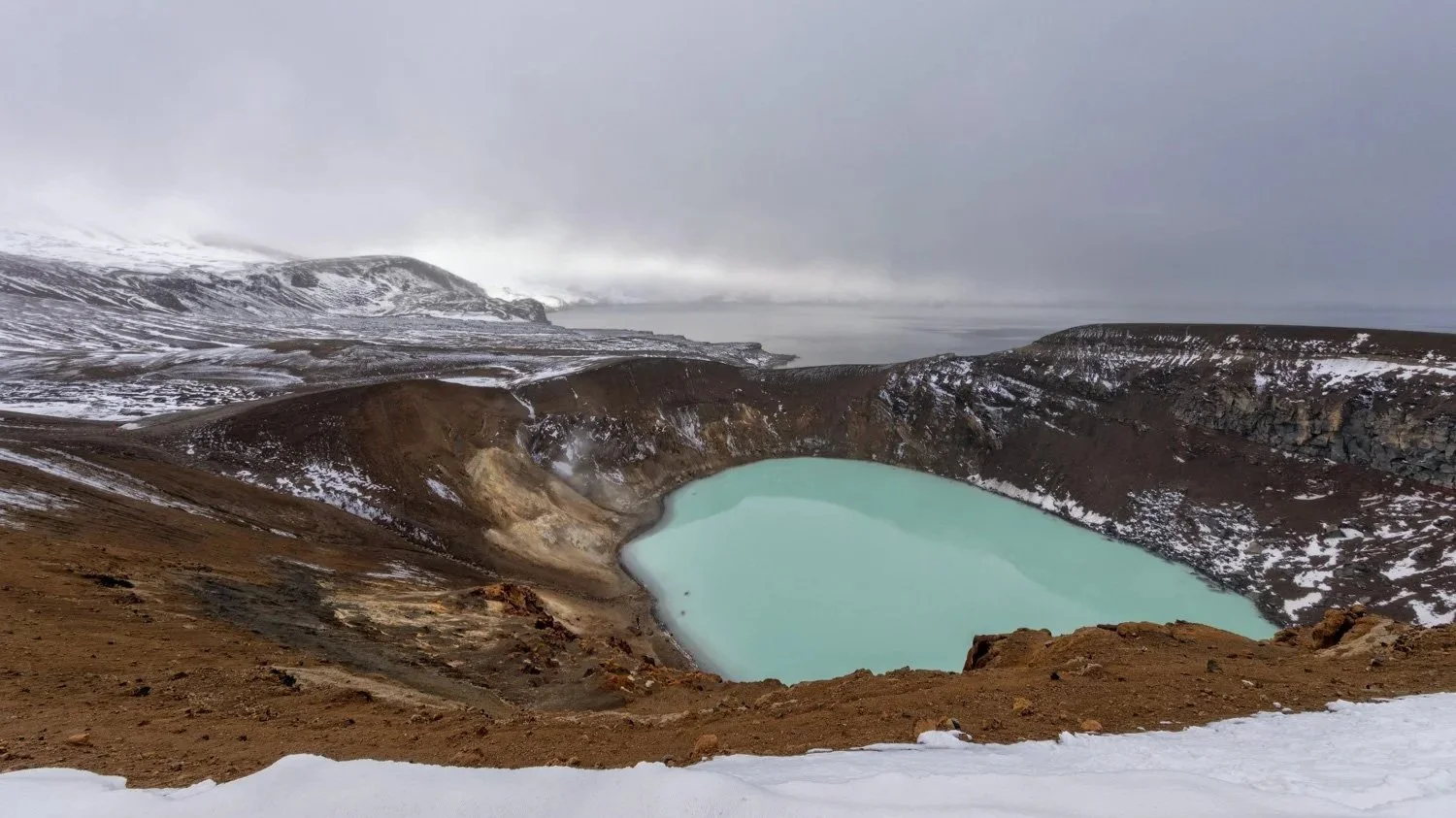 Vulkansee in einer caldera, umgeben von schneebedeckten Hügeln und dunklem vulkanischem Gestein, bei bewölktem Himmel.