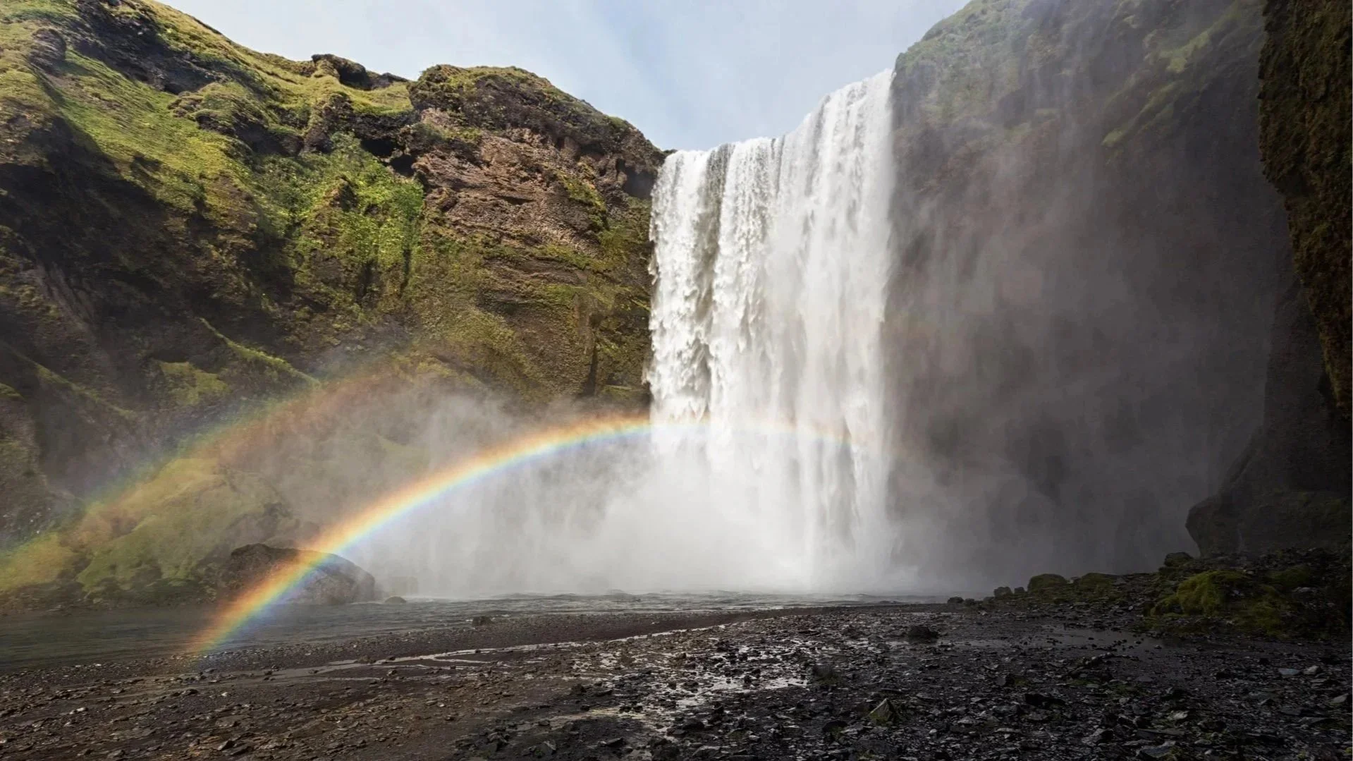 Ein Wasserfall fließt in eine Schlucht, mit einem Regenbogen im Wassernebel im Vordergrund.