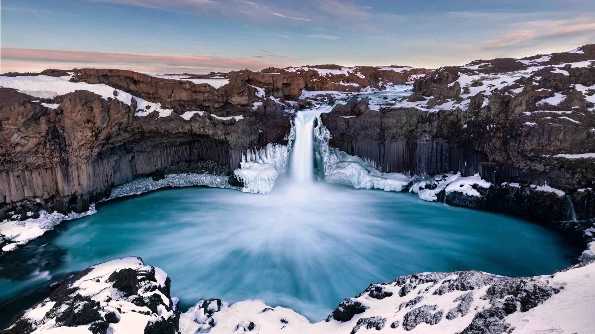 Ein Wasserfall in einer winterlichen Landschaft mit schneebedeckten Felsen und gefrorenem Wasser, unter einem Himmel mit blauen und pinkfarbenen Wolken.