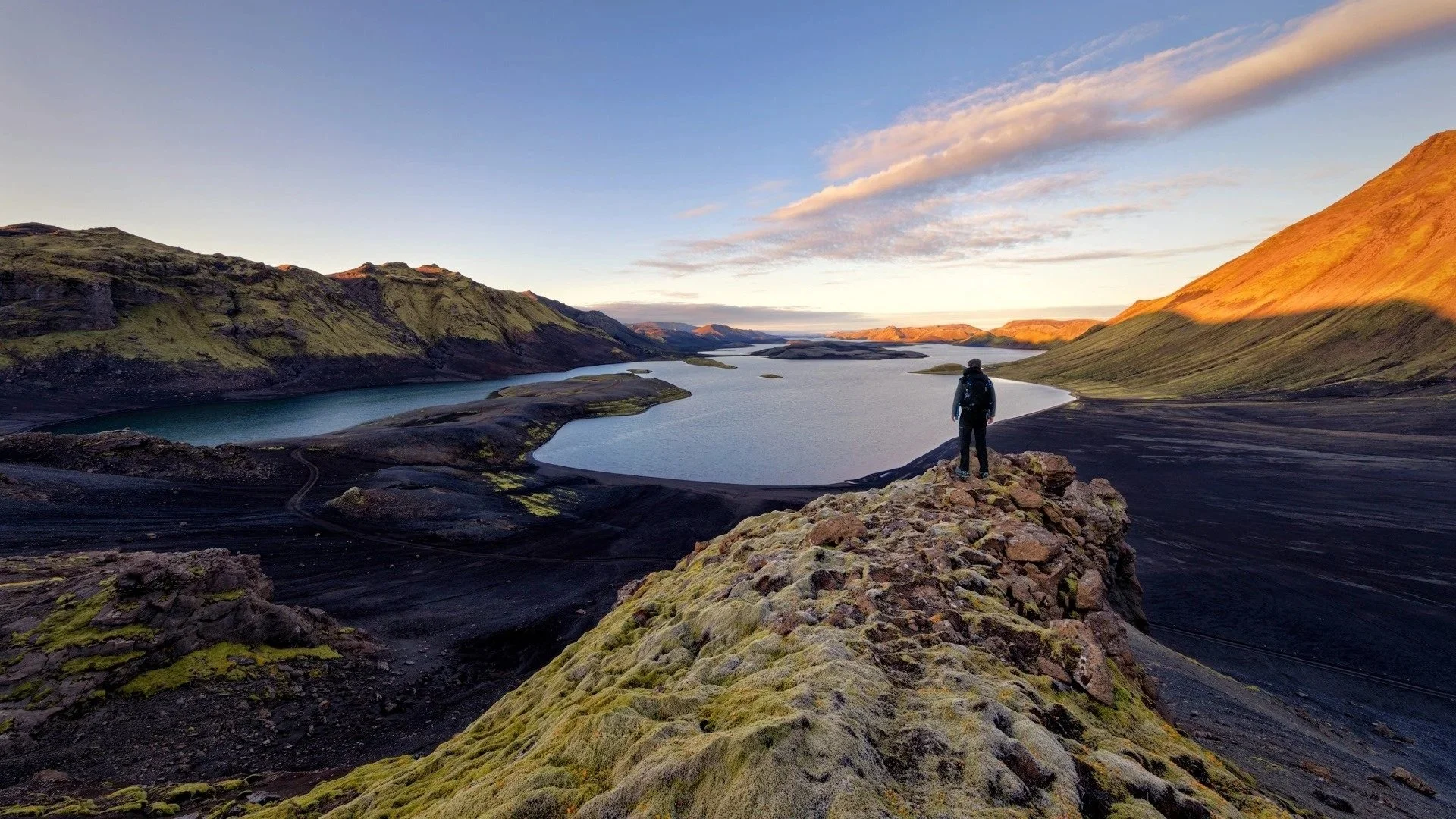 Ein einzelner Wanderer mit Rucksack steht auf einem Felsen und blickt auf einen malerischen See, umgeben von grünen Hügeln und Bergen bei Sonnenuntergang.