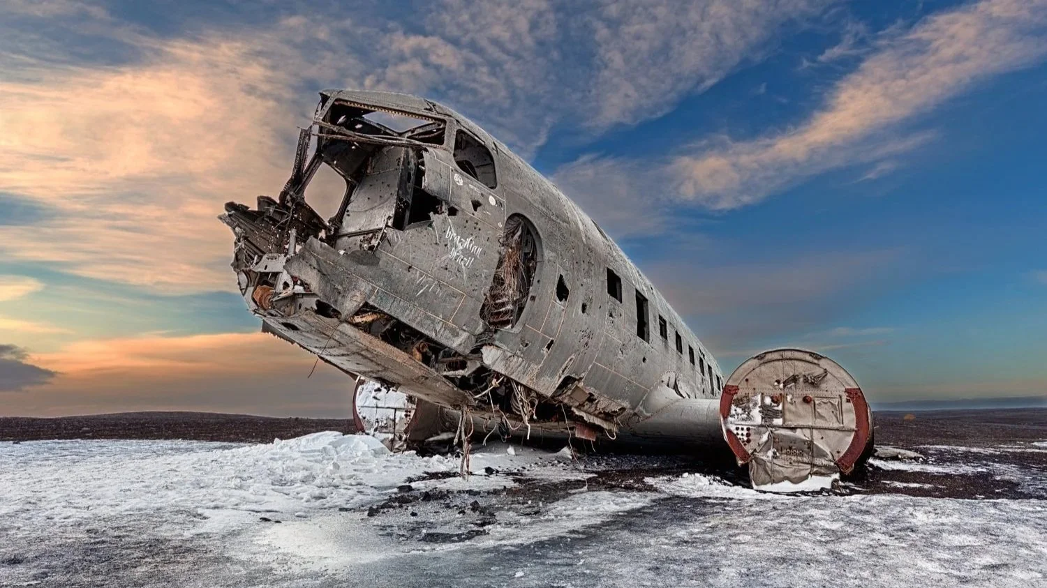 Ein abgestürztes Flugzeug liegt im Schnee auf einer kargen, eisigen Landschaft bei Sonnenuntergang, mit einem dramatischen Himmel im Hintergrund.